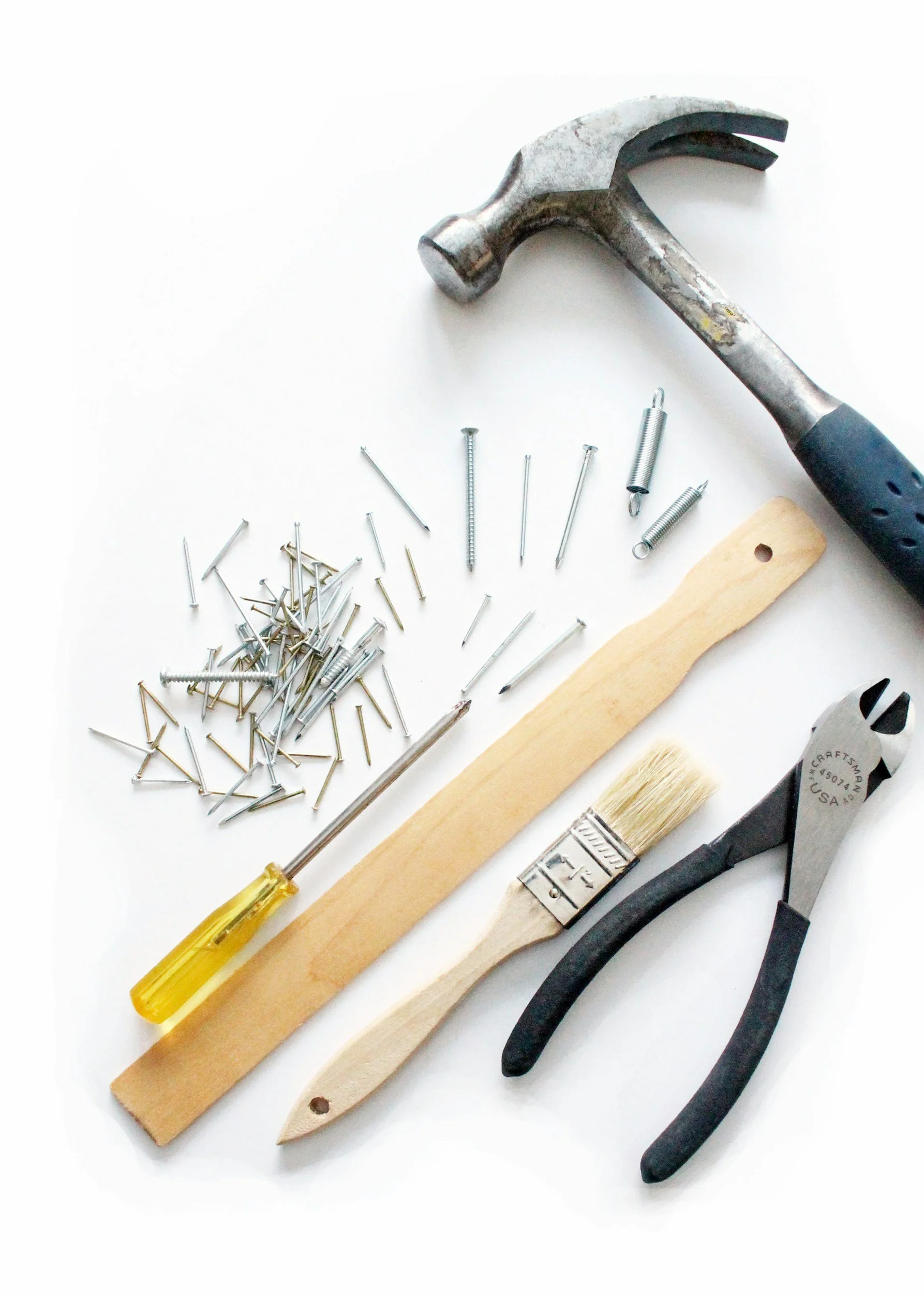 A collection of hand tools including a hammer, pliers, a screwdriver, a wooden paint stir stick, a paintbrush, a large number of nails, and some small springs, all arranged on a white surface.