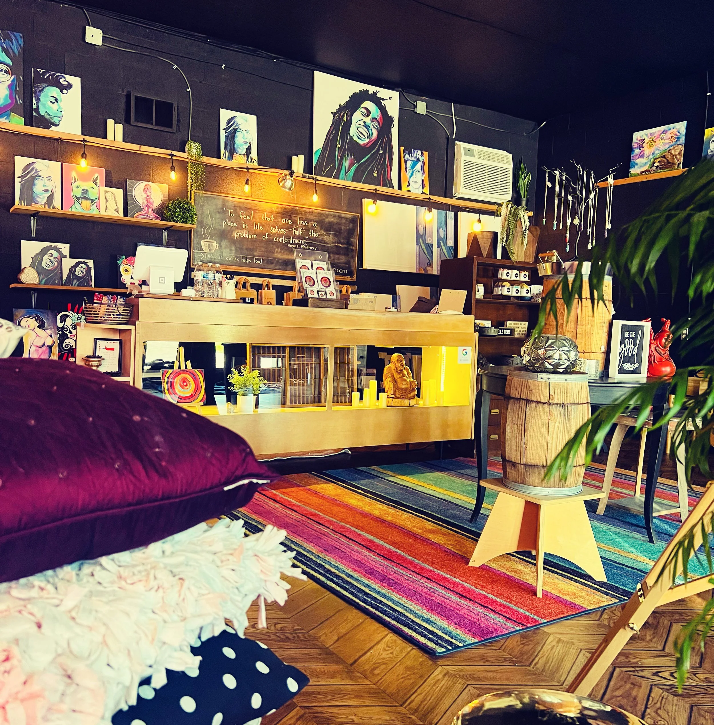 Art studio or creative workspace with colorful paintings on dark walls, a wooden counter with art supplies and decorations, a vibrant striped rug on wooden floor, a large leafy plant in a wooden barrel planter, and cozy seating and tables. Warm lighting highlights the eclectic decor.