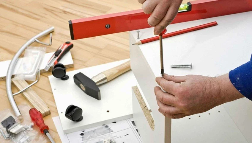 Person assembling furniture with a screwdriver, surrounded by tools and hardware on a wooden floor.