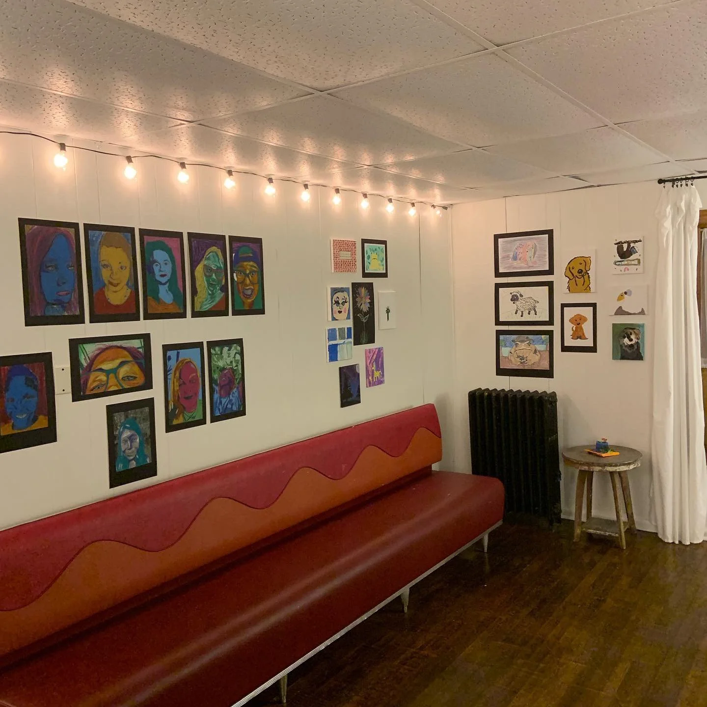 A room with white walls decorated with various colorful artwork, including portraits, animals, and abstract designs. A string of warm lights hangs across the ceiling. There's a long red and orange bench along the wall, a small round wooden table with a few items, a black radiator, and a white curtain near a window.