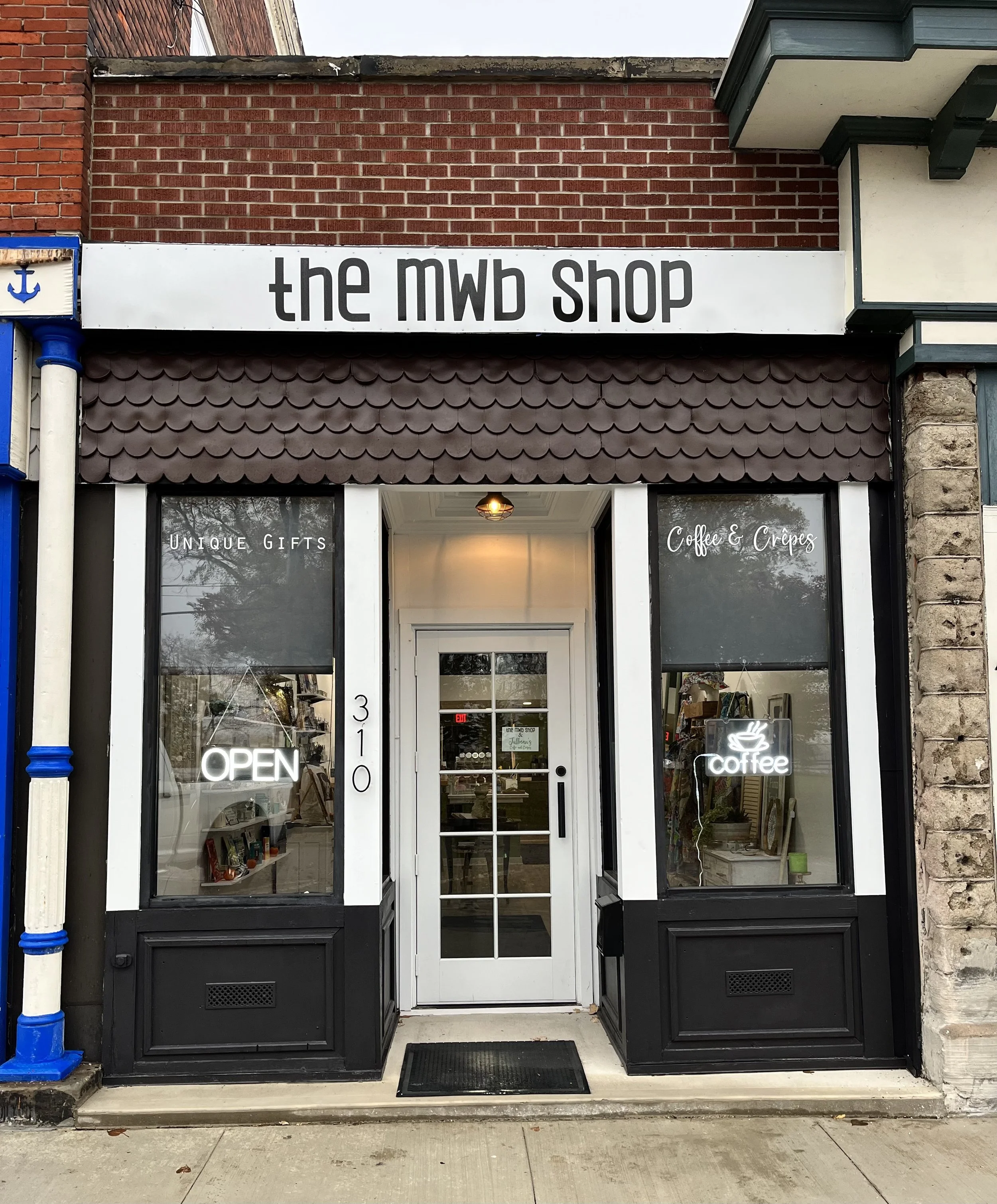 The storefront of 'the mwb shop' featuring black and white exterior, signs for 'Unique Gifts' and 'Coffee & Crêpes,' neon signs that say 'OPEN' and 'coffee,' a white door with glass panels, and large windows on each side displaying interior shelves.