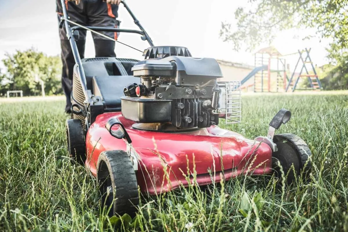 Close-up of a red lawnmower on grass with a person standing behind it, in a park or yard with trees and playground equipment in the background.