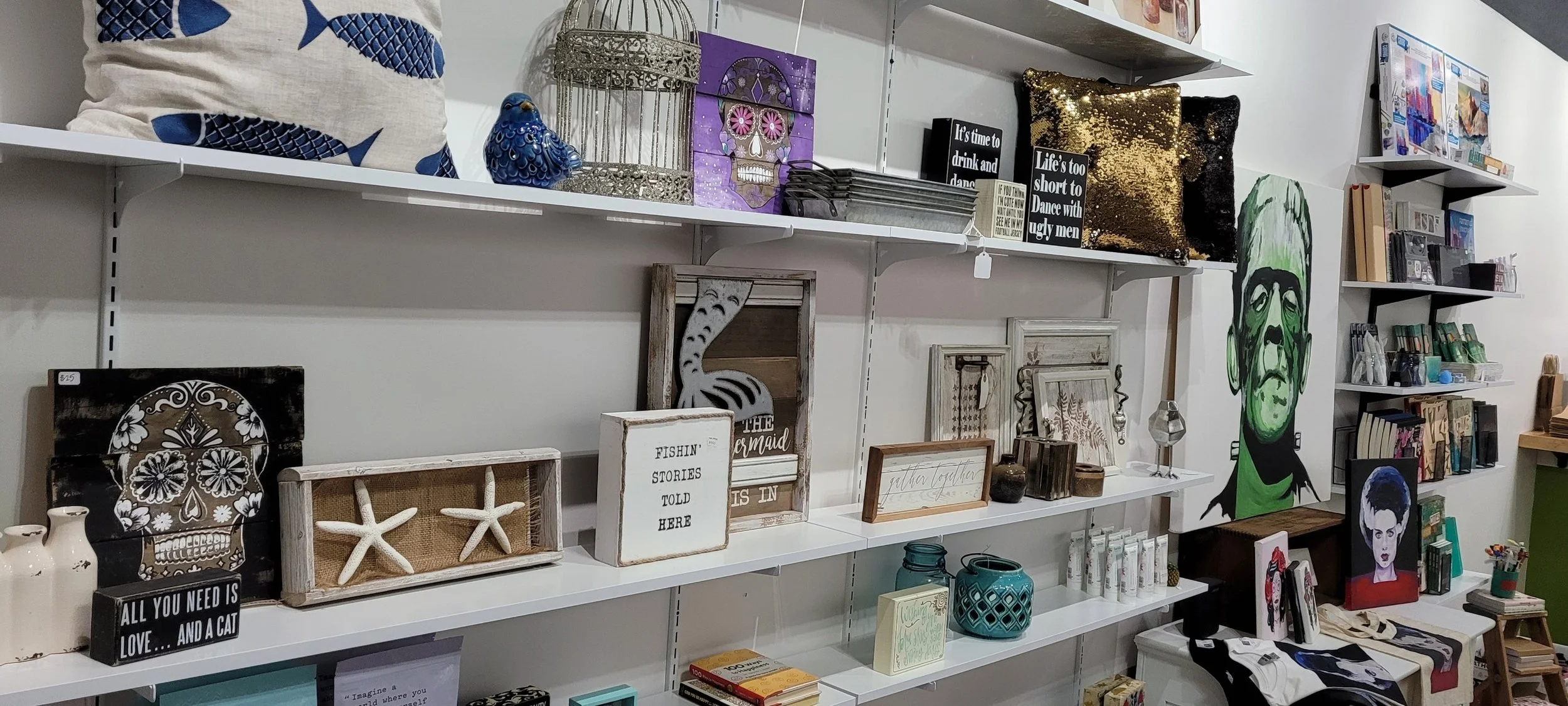 Wall display with various decorative items including framed art, pillows, sculptures, candles, and books, in a store setting.