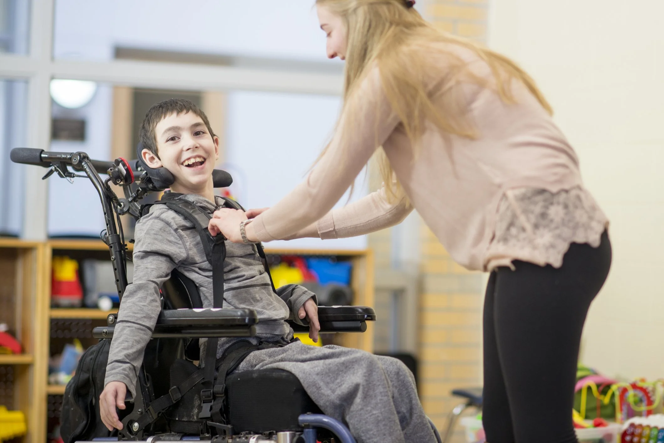 A young boy in a motorized wheelchair smiling while a woman adjusts his shirt in a room with shelves of toys and a window.