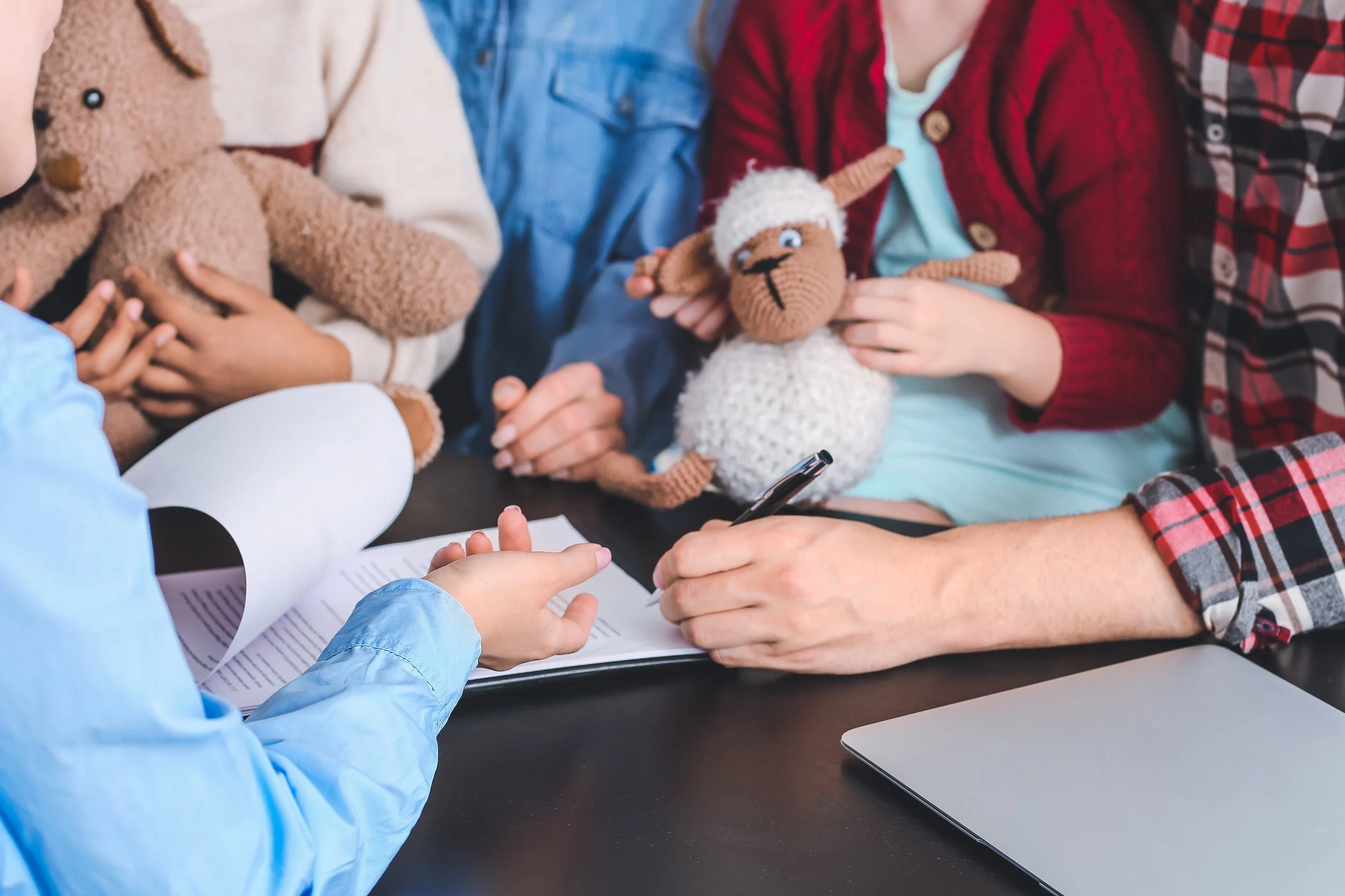 A person in a blue jacket is sitting at a table with others, holding a black pen and writing or signing a document. Several children are sitting around the table, each holding or playing with plush sheep toys and teddy bears, in a cozy setting.