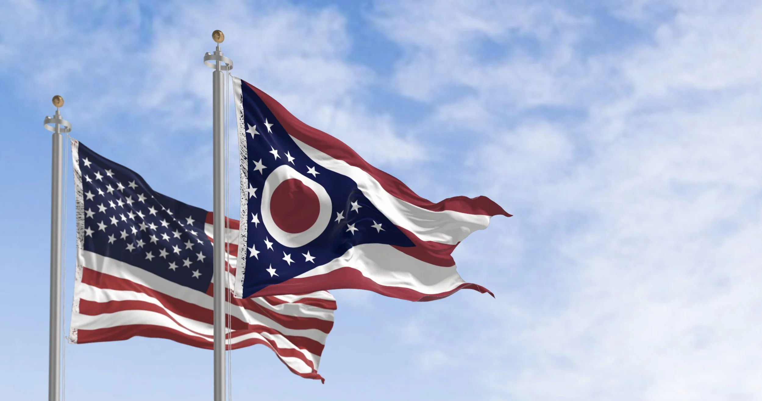 Two American flags and one Ohio state flag flying on flagpoles against a blue sky with scattered clouds.