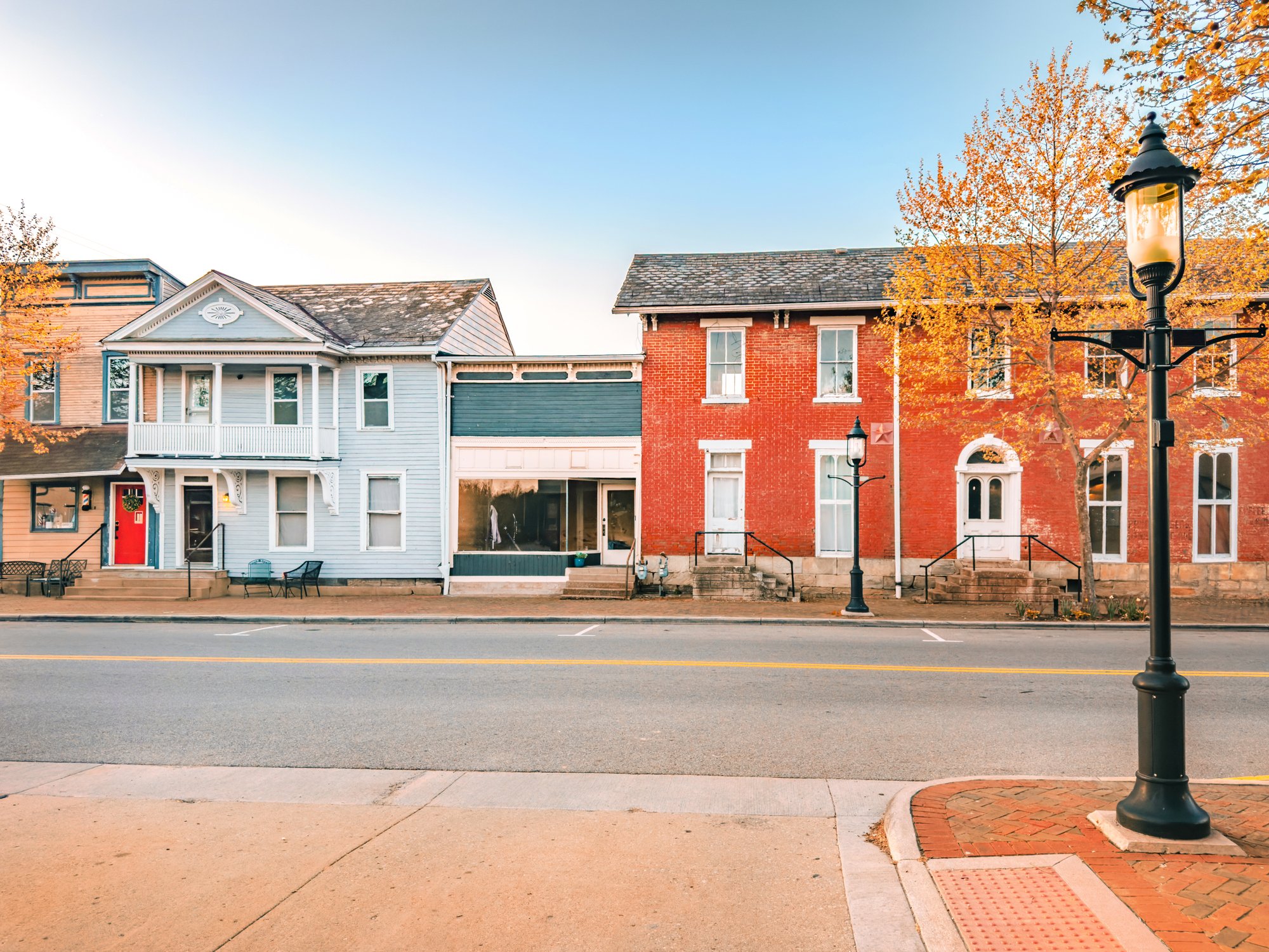A row of three houses on a quiet street during autumn. The house on the left is light blue with a red door, the middle house has a storefront style with large window, and the house on the right is red brick. The street has street lamps and trees with orange leaves, and the sky is clear.