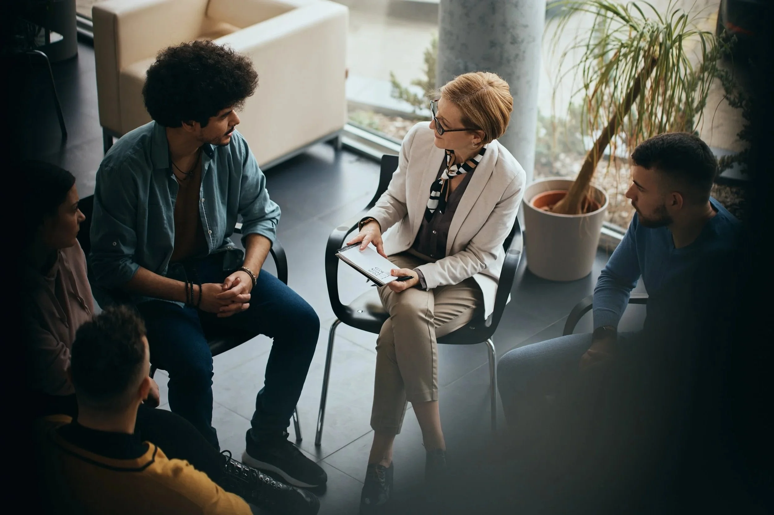 A woman with red hair and glasses is sitting in a chair, holding a notebook, and speaking with four young adults in a modern office or lounge area, near a large window with a potted plant in the background.