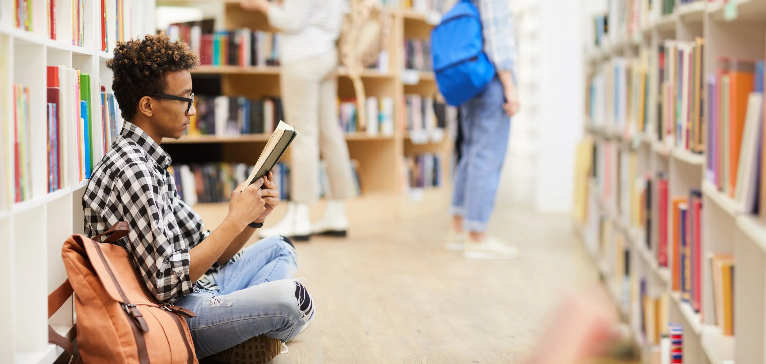 A person with curly hair, glasses, and a checkered shirt sitting on the floor reading a book in a library, with a backpack beside them. Other people in the background are browsing bookshelves.