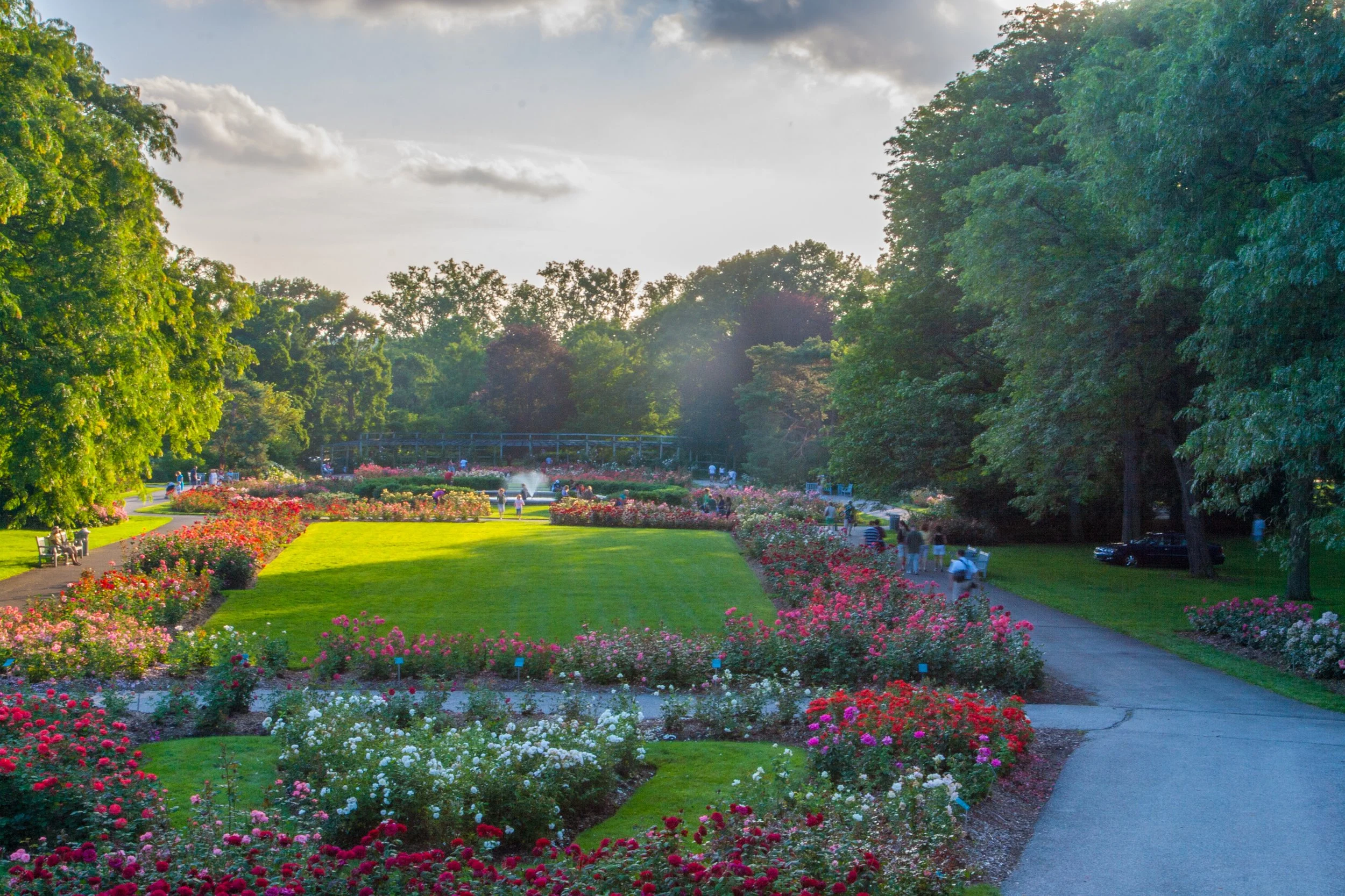 A scenic park with vibrant flower beds, a well-maintained grass lawn, and walking paths. People are strolling along the paths, enjoying the lush greenery and blooming flowers under a partly cloudy sky.