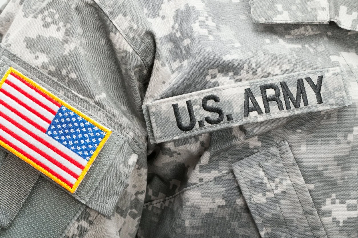 Close-up of a U.S. Army uniform with a gray camouflage pattern, showing a United States flag patch on the sleeve and a 'U.S. ARMY' name tape above the chest.