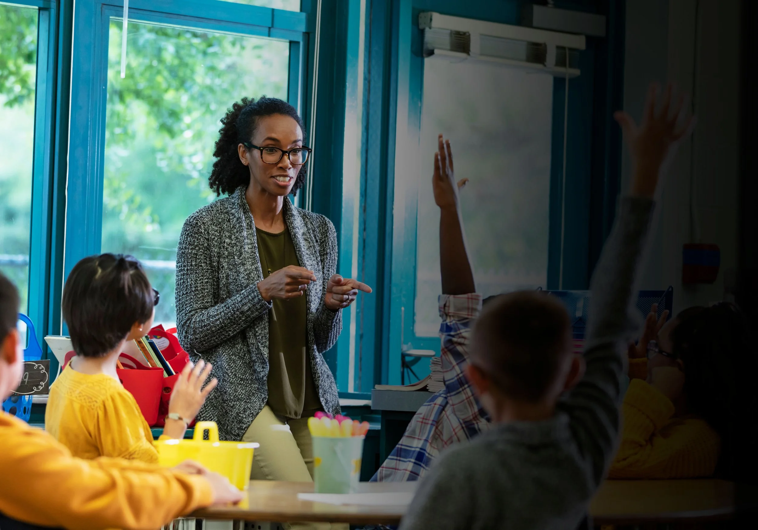 A woman teacher in a classroom interacting with young students, some of whom have their hands raised, during a lesson or discussion.