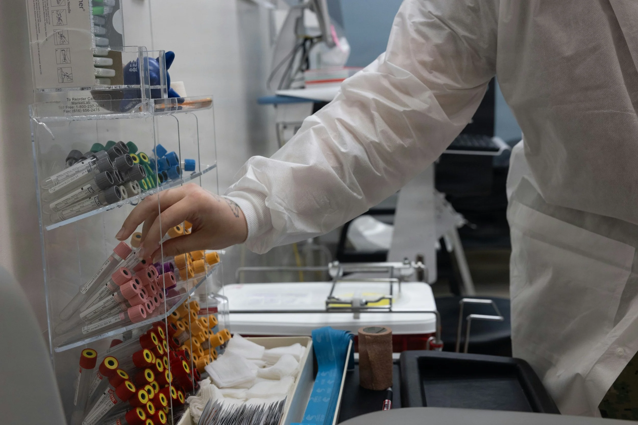 A person in medical protective clothing reaching for blood collection tubes in a laboratory setting with various lab supplies around.