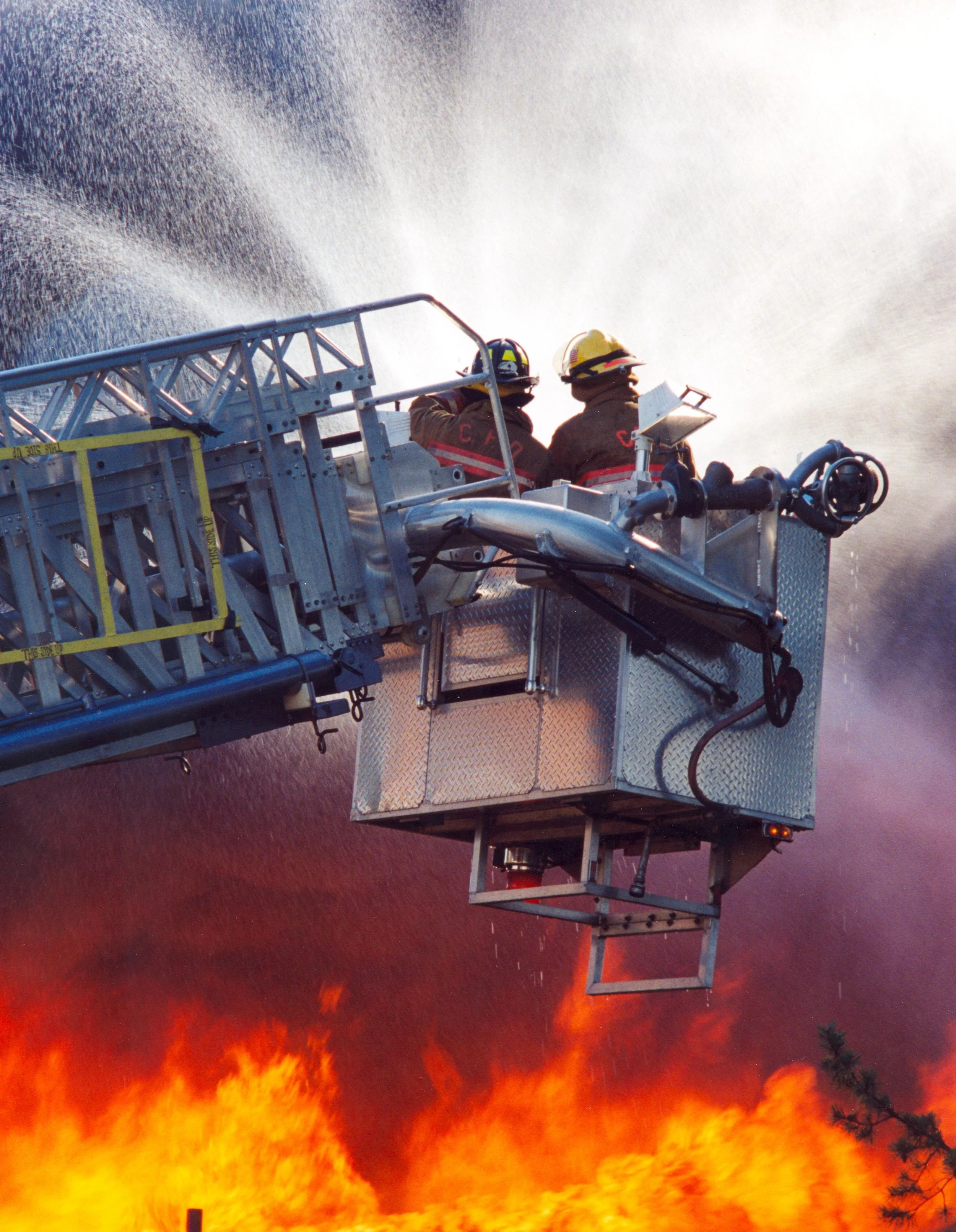 Two firefighters in protective gear and helmets on a hydraulic fire truck extend basket over large flames and smoke during a fire.