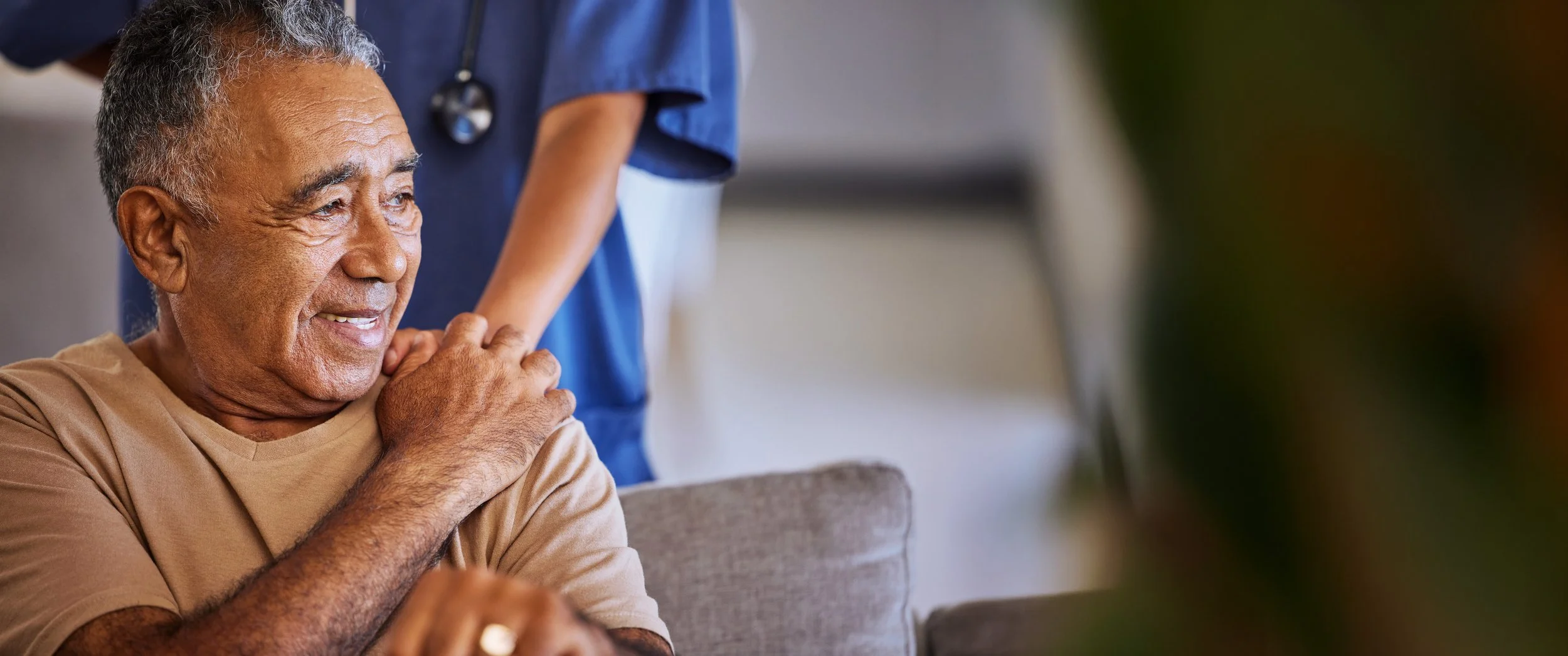 An elderly man smiling and sitting on a couch in a healthcare setting, with a healthcare professional in blue scrubs and a stethoscope behind him.