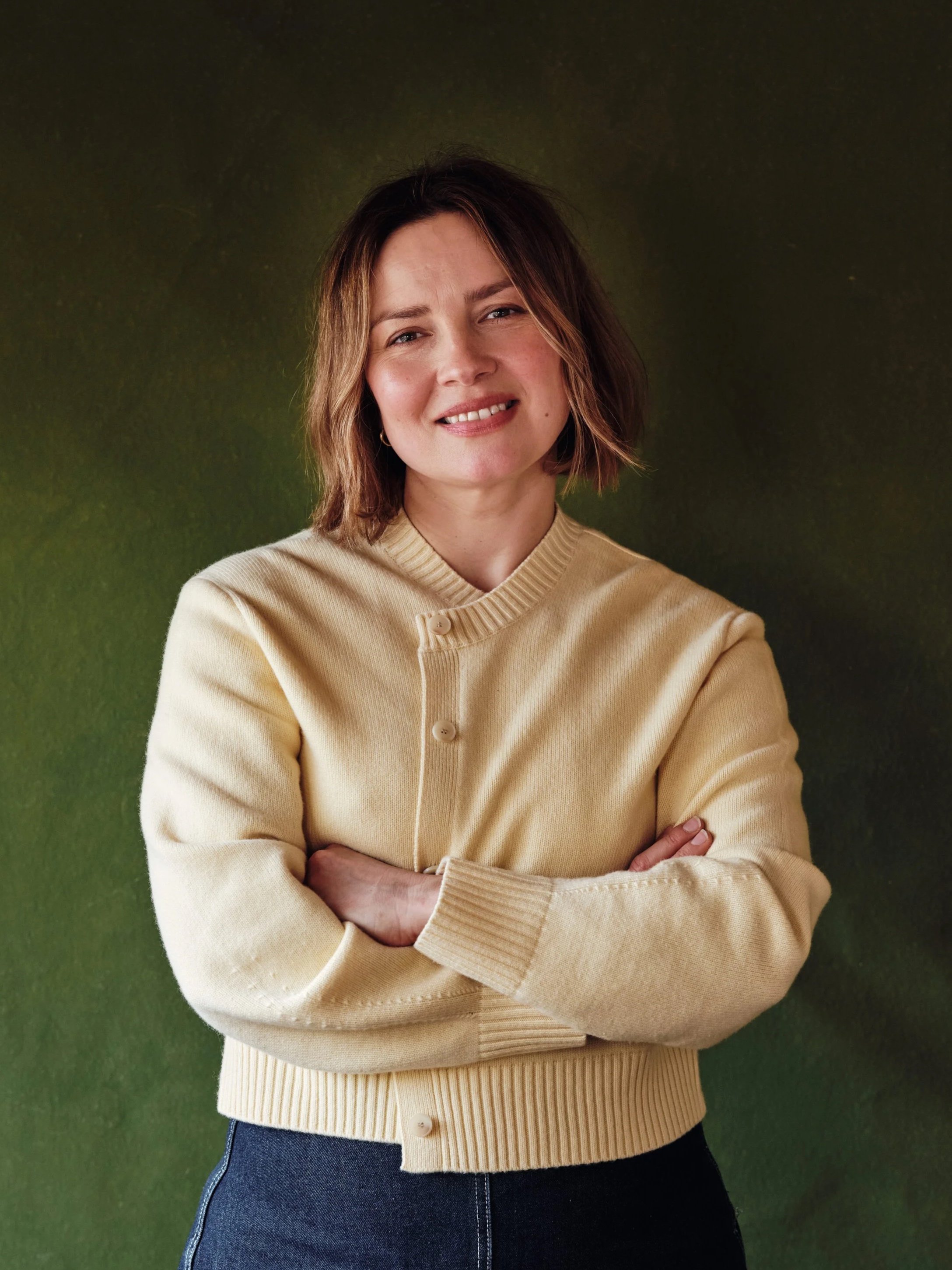 Ellie Stidolph, a personal stylist with short brown hair smiling, wearing a butter yellow cardigan with her arms crossed, standing against a green background.