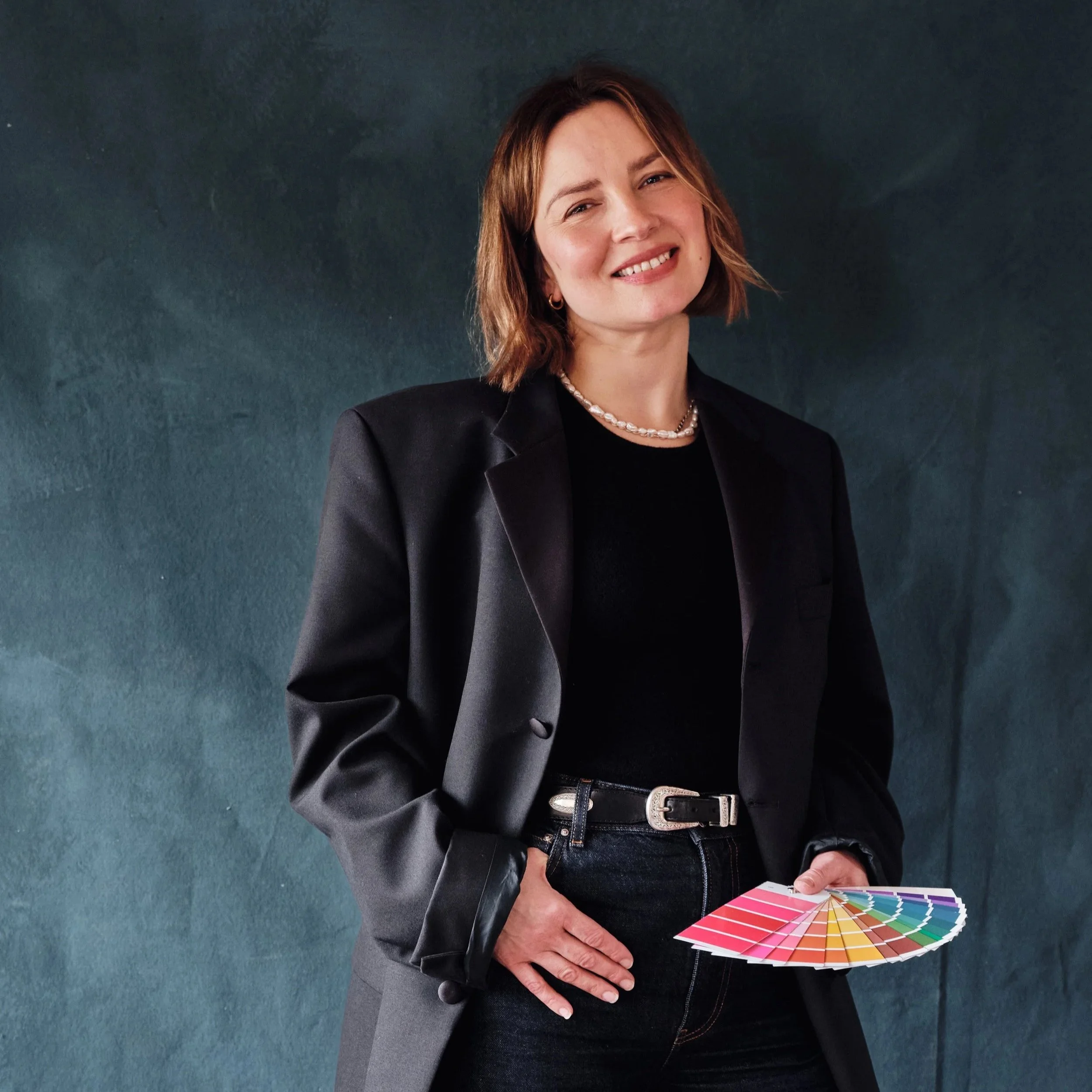 Ellie Stidolph, a stylist  in a black blazer and jeans holding a fan of colour analysis swatches, standing against a dark green wall, smiling at the camera.