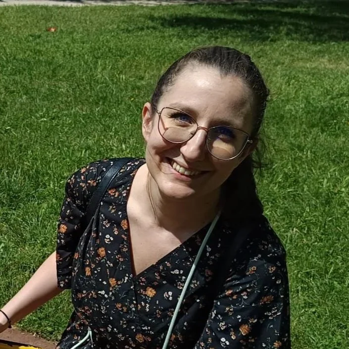 Femme souriante portant des lunettes, vêtue d'une robe noire à motifs floraux, assise sur l'herbe dans un parc ensoleillé.