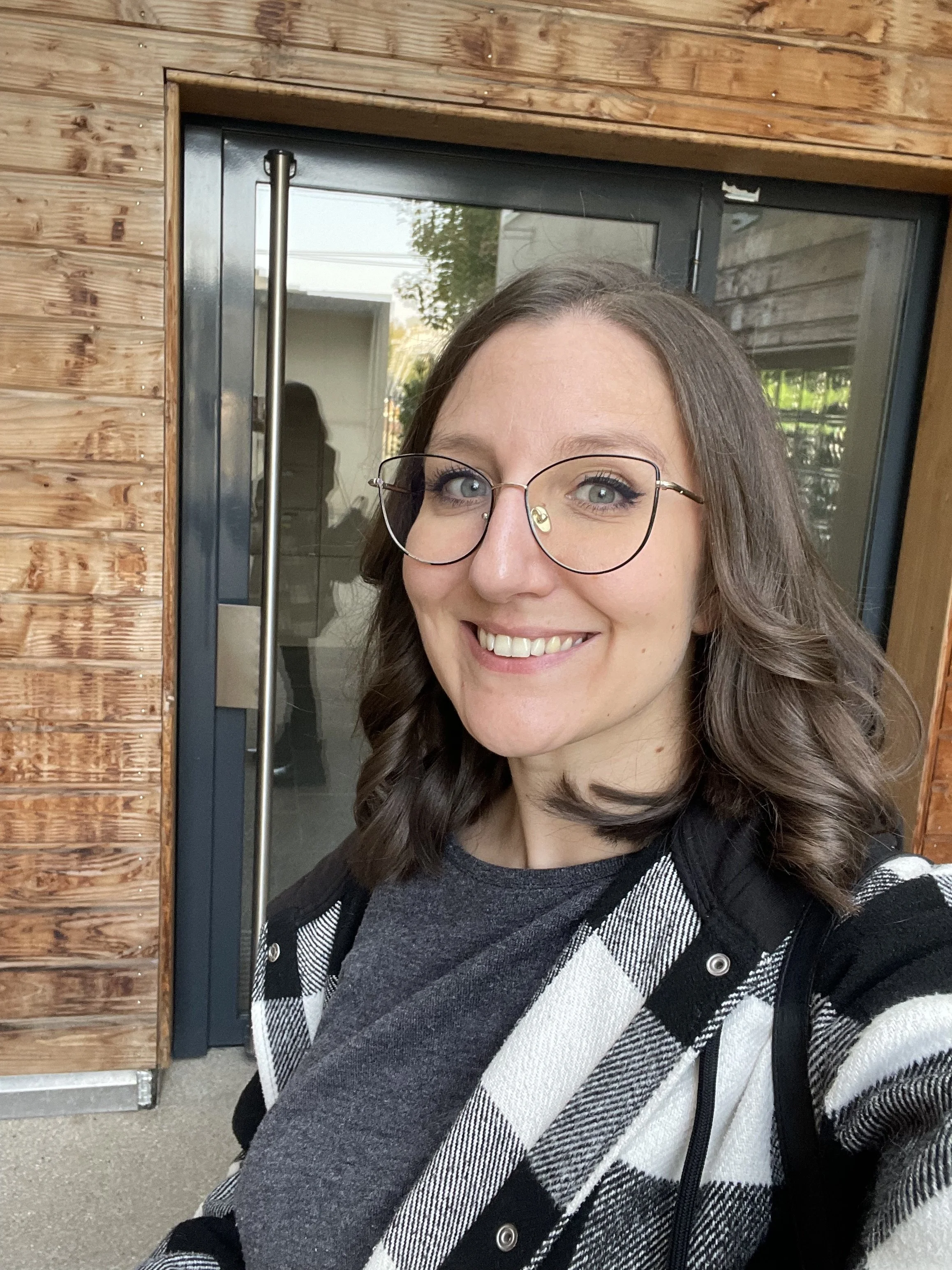 Une femme souriante portant des lunettes, écharpe à carreaux noir et blanc, en extérieur devant une porte en verre avec un cadre noir, et un mur en bois rustique derrière elle.