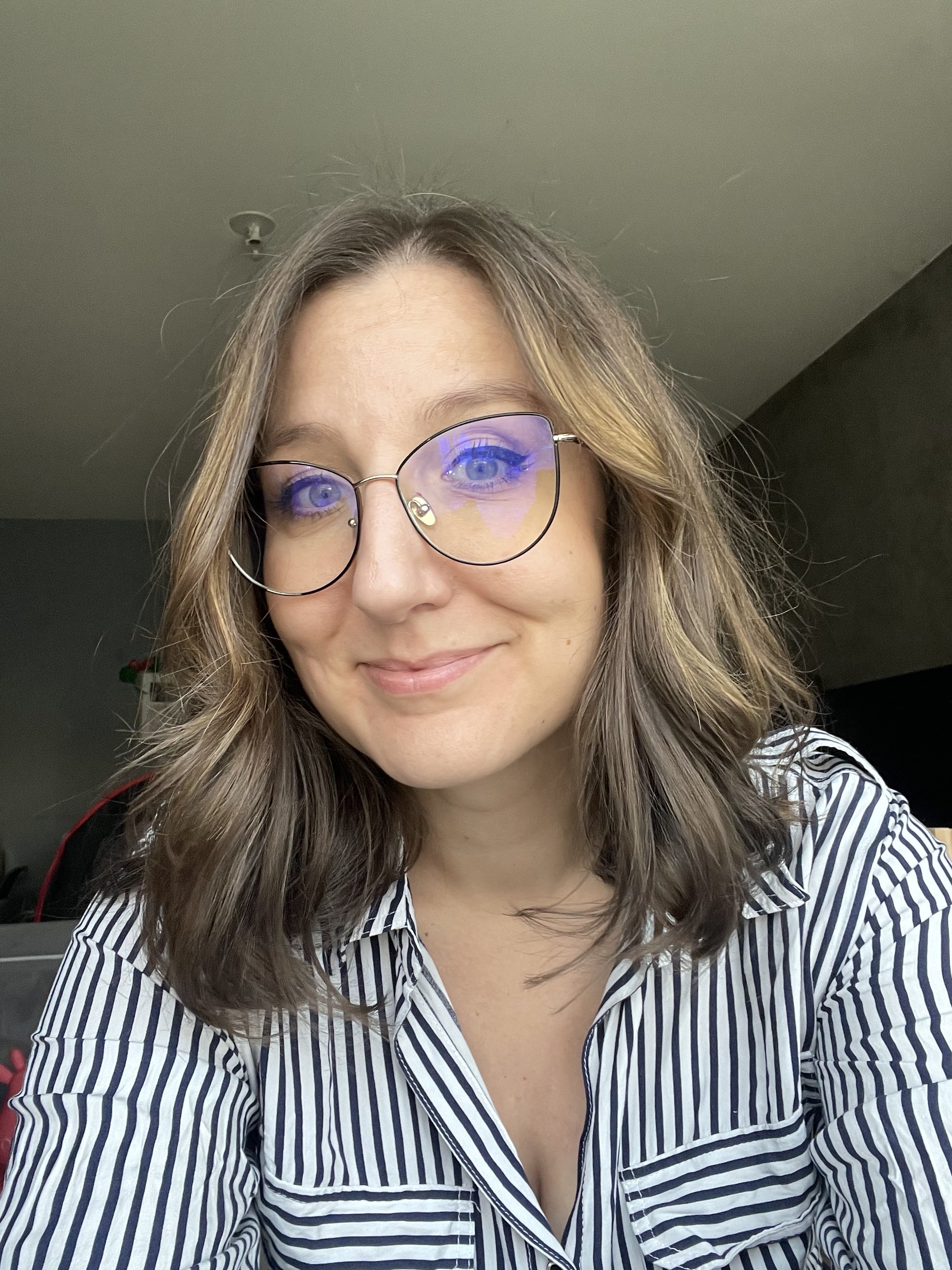 Une femme avec des cheveux bruns ondulés portant des lunettes rondes et un chemisier à rayures bleues et blanches, souriant à la caméra, à l'intérieur.