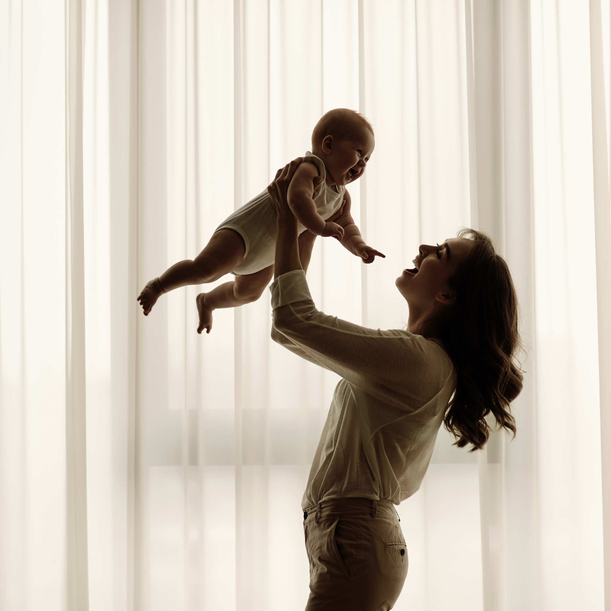 Une femme souriante lève un bébé dans une pièce avec des rideaux blancs en arrière-plan, créant une silhouette contre la lumière naturelle.