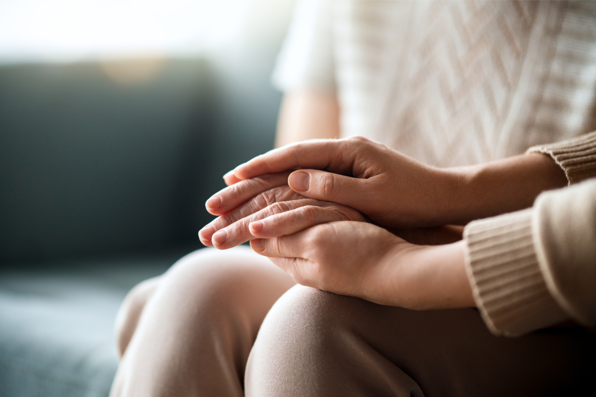 Deux mains qui se tiennent, posées l'une sur l'autre, sur les genoux d'une personne assise dans un intérieur lumineux.
