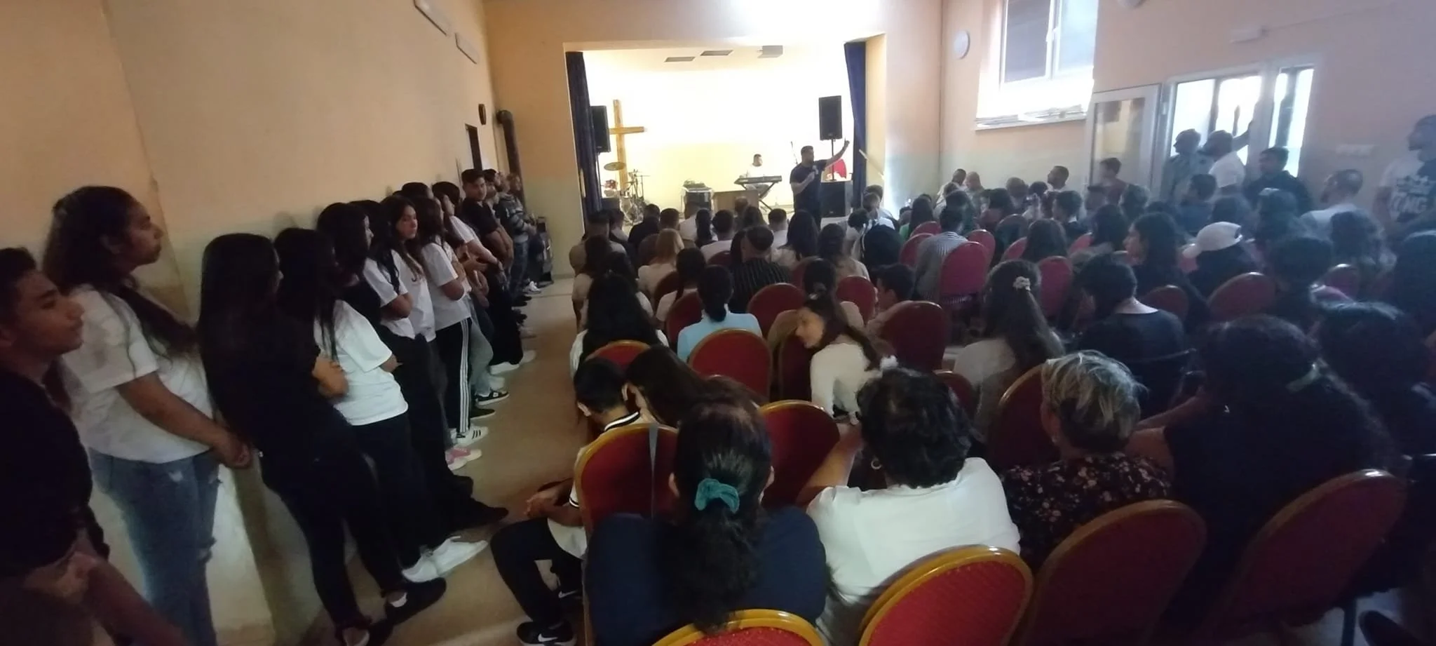 A crowded indoor gathering with a stage at the front, where a person is speaking or singing into a microphone. Attendees are seated and standing, watching the stage. The room has walls with large windows and a religious cross is visible at the front,