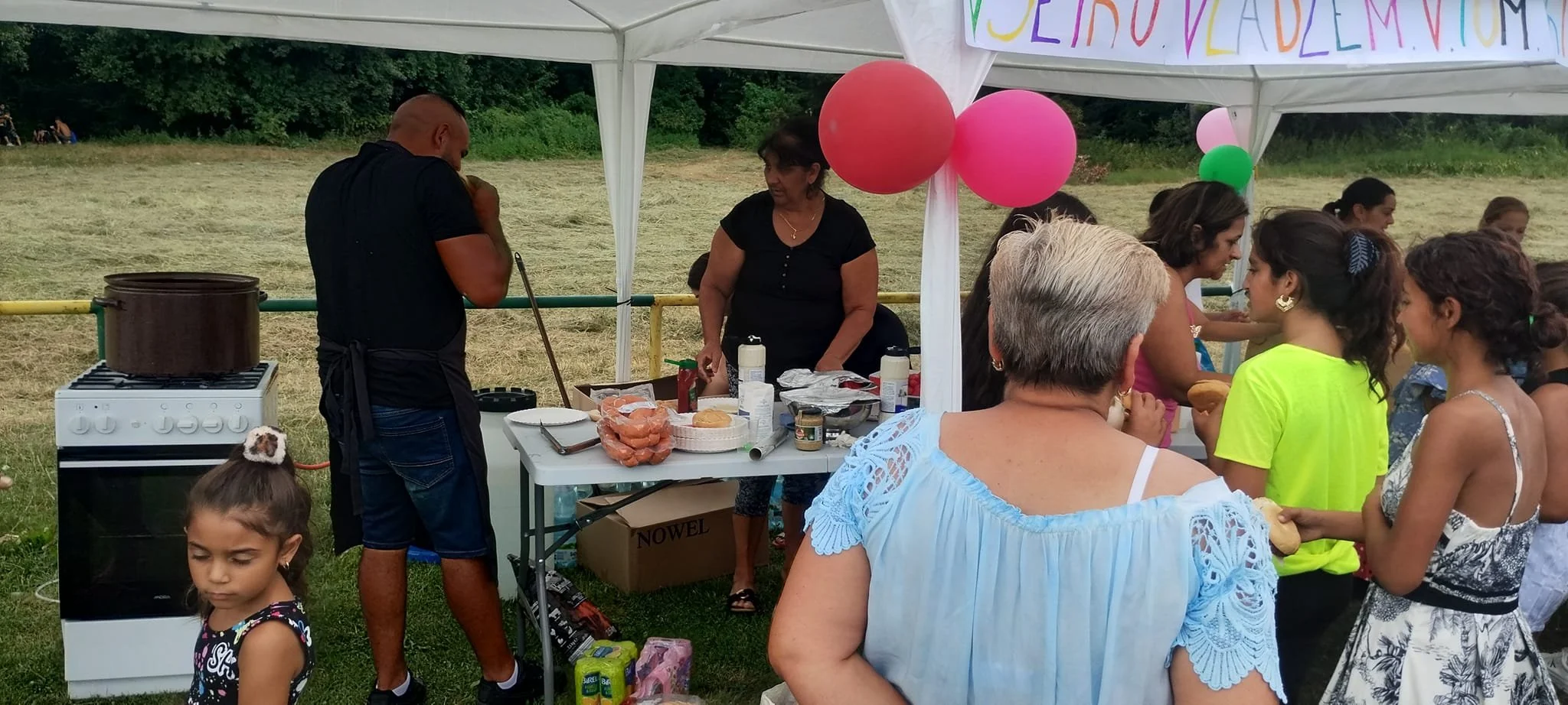 People gathered at a food stall at an outdoor event under a white canopy decorated with pink and green balloons. A woman is preparing food behind the stall, and children and adults are in line, some holding food.