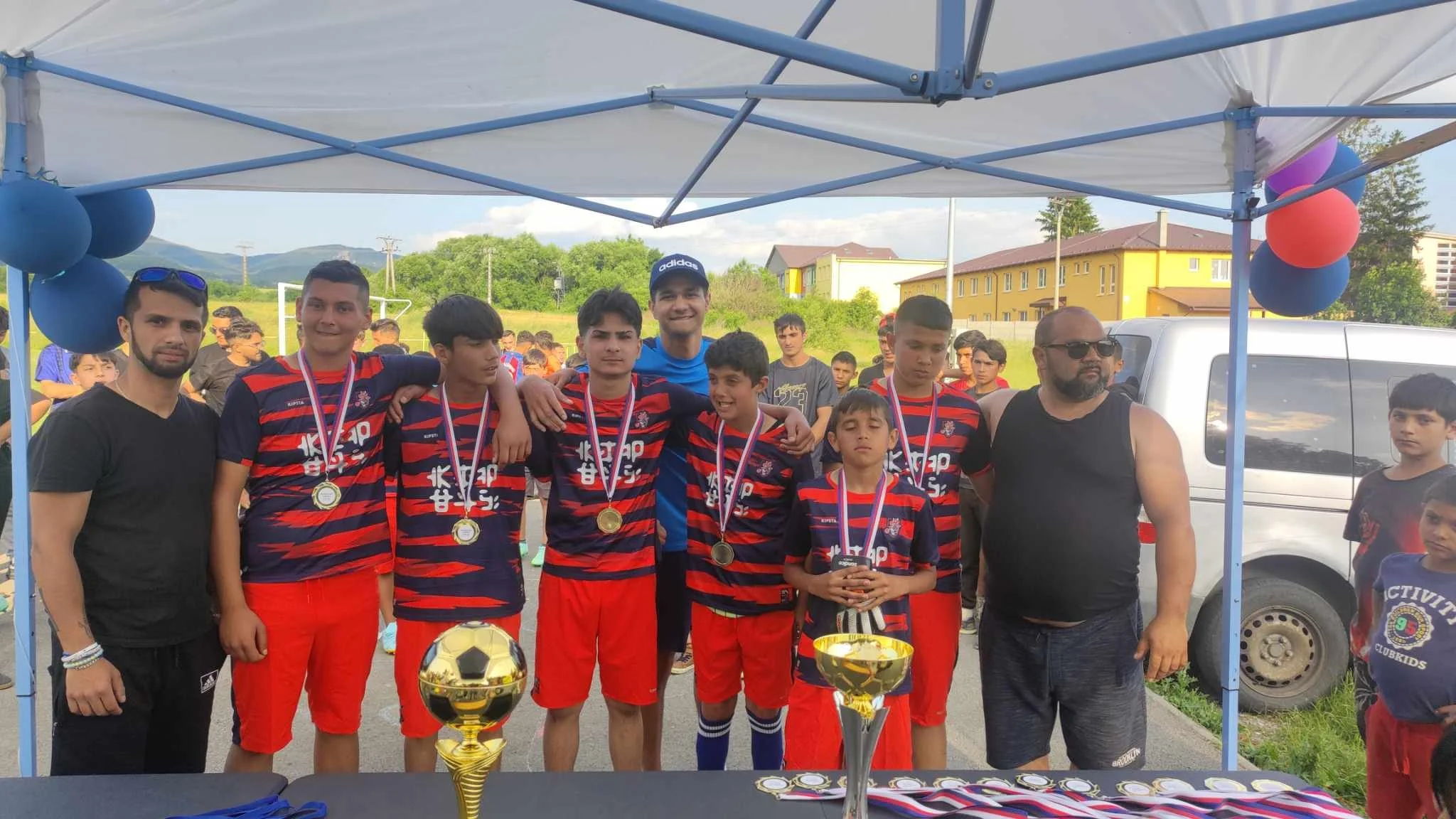 A group of young soccer players with medals posing for a photo under a tent, with trophies in front and a crowd in the background, celebrating their victory.