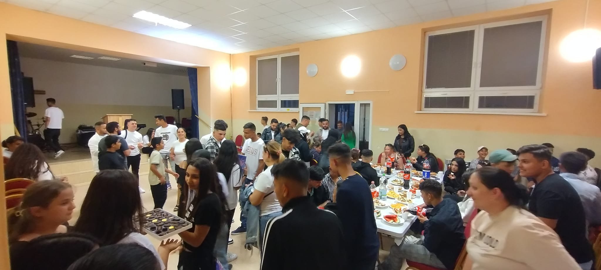 A large indoor gathering or celebration with people socializing, eating, and serving food. Some individuals are sitting at a long table with drinks and snacks, while others are standing and mingling.