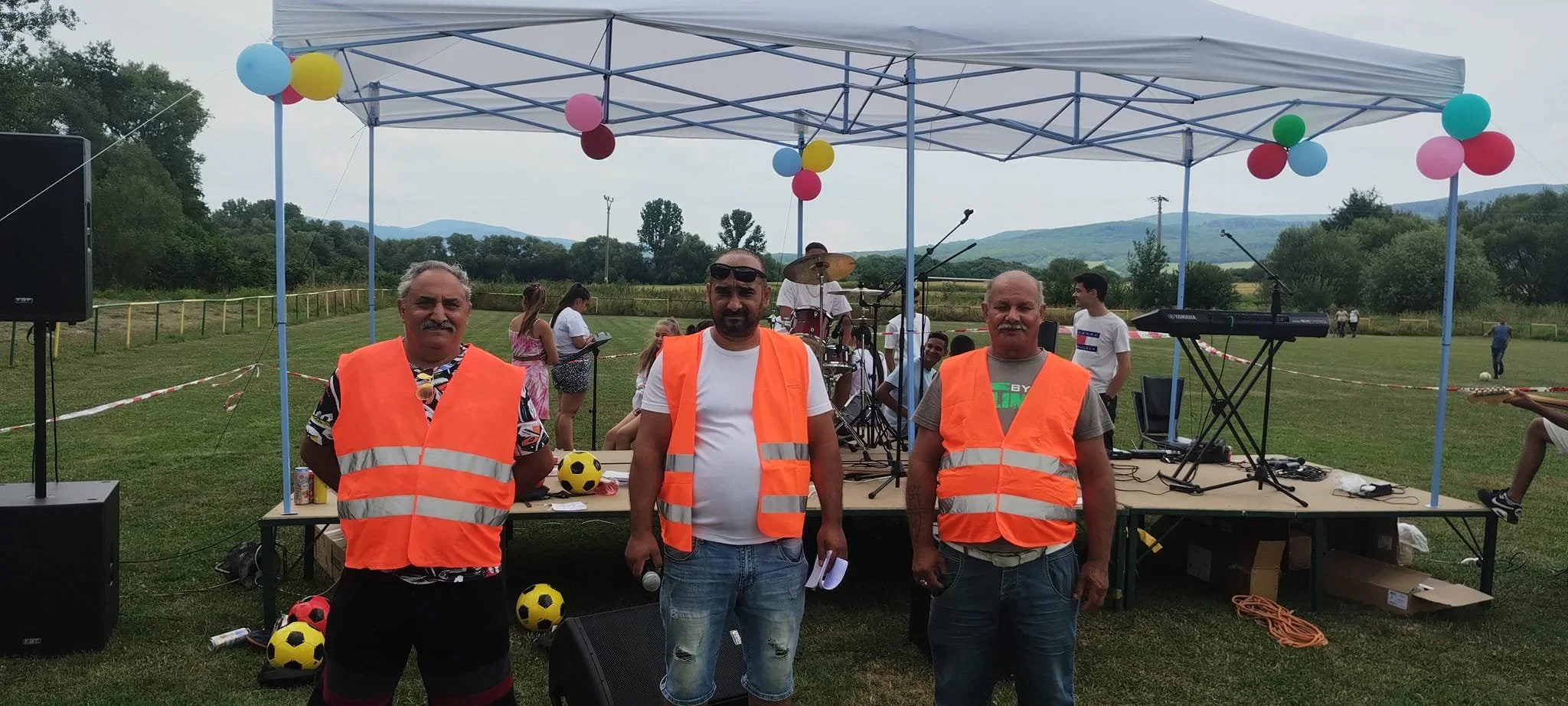 Outdoor event with a stage under a canopy decorated with balloons, featuring three men wearing orange reflective safety vests standing in front of the stage, which has musical instruments and microphones. Several people and children are in the backgr