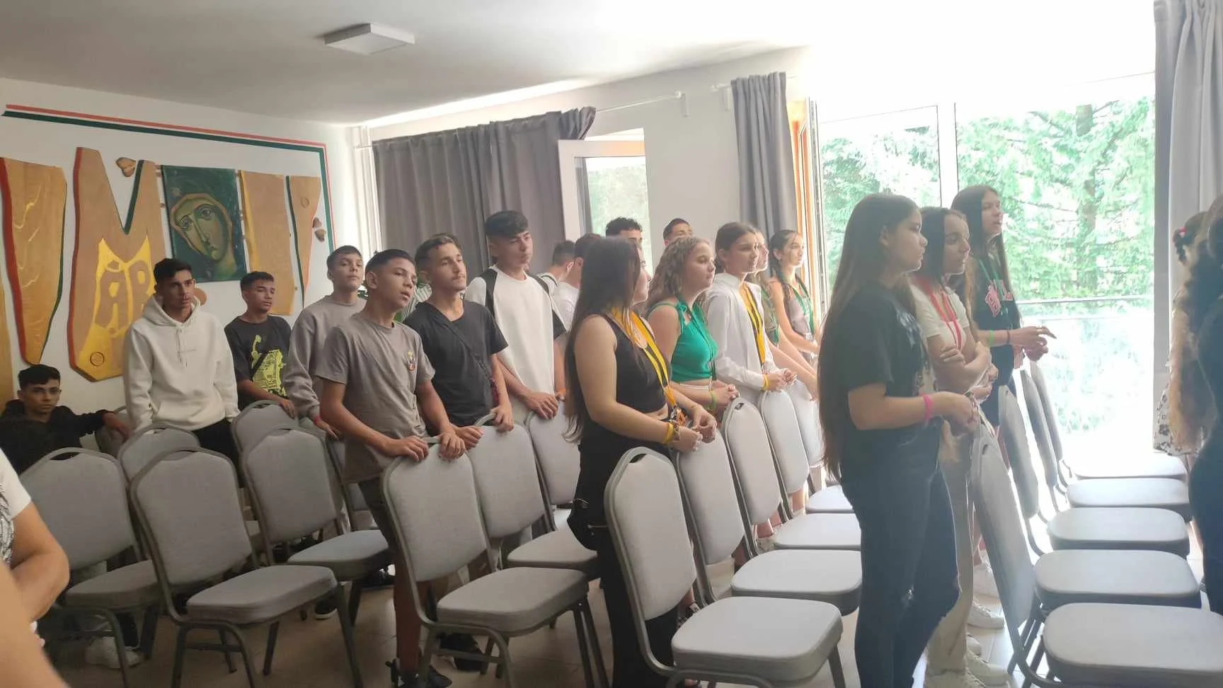 Group of teenagers standing inside a room, some near the open door, with chairs arranged in rows, and a colorful mural on the wall.