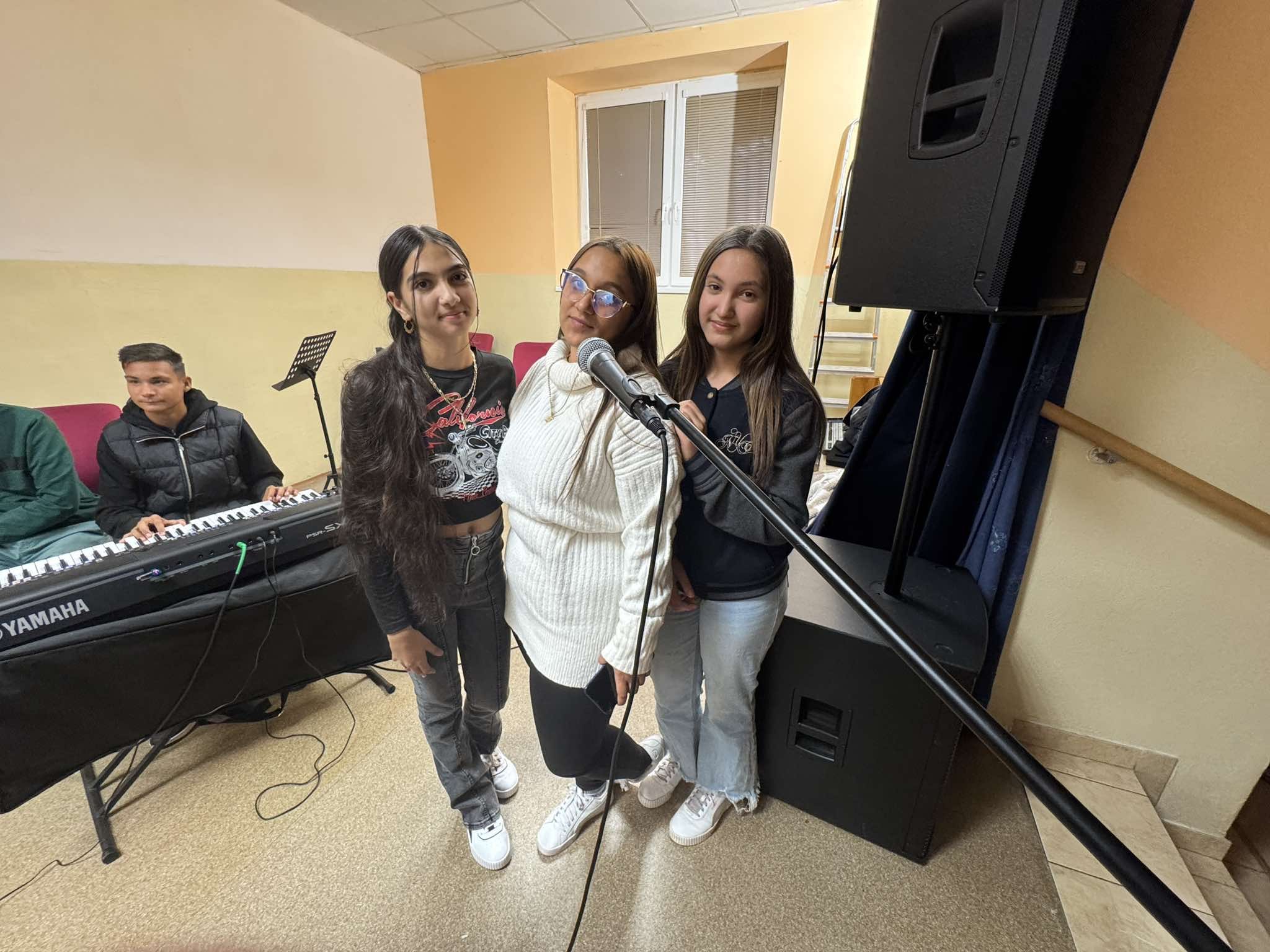 Group of four young people, three females and one male, posing in a room with musical equipment, including a keyboard and microphone.