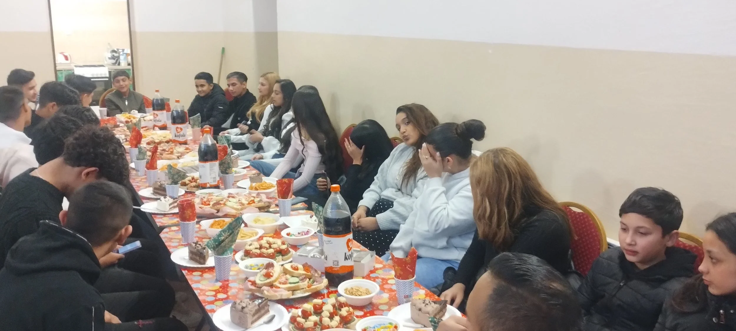 Group of young people gathered around a banquet table with various foods and drinks, at a holiday or celebration event.
