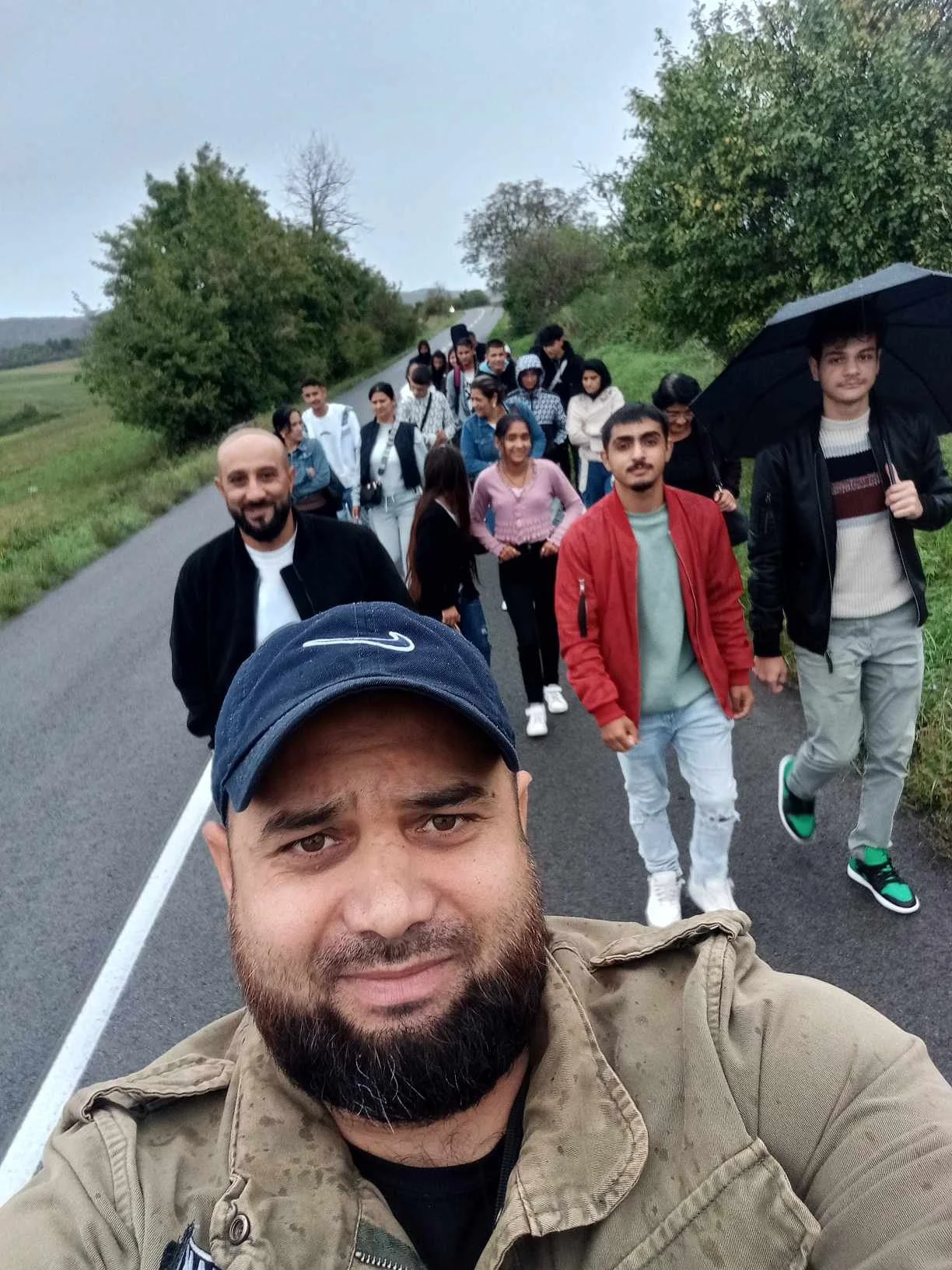 A man taking a selfie with a group of people walking on a rural road during a cloudy day.