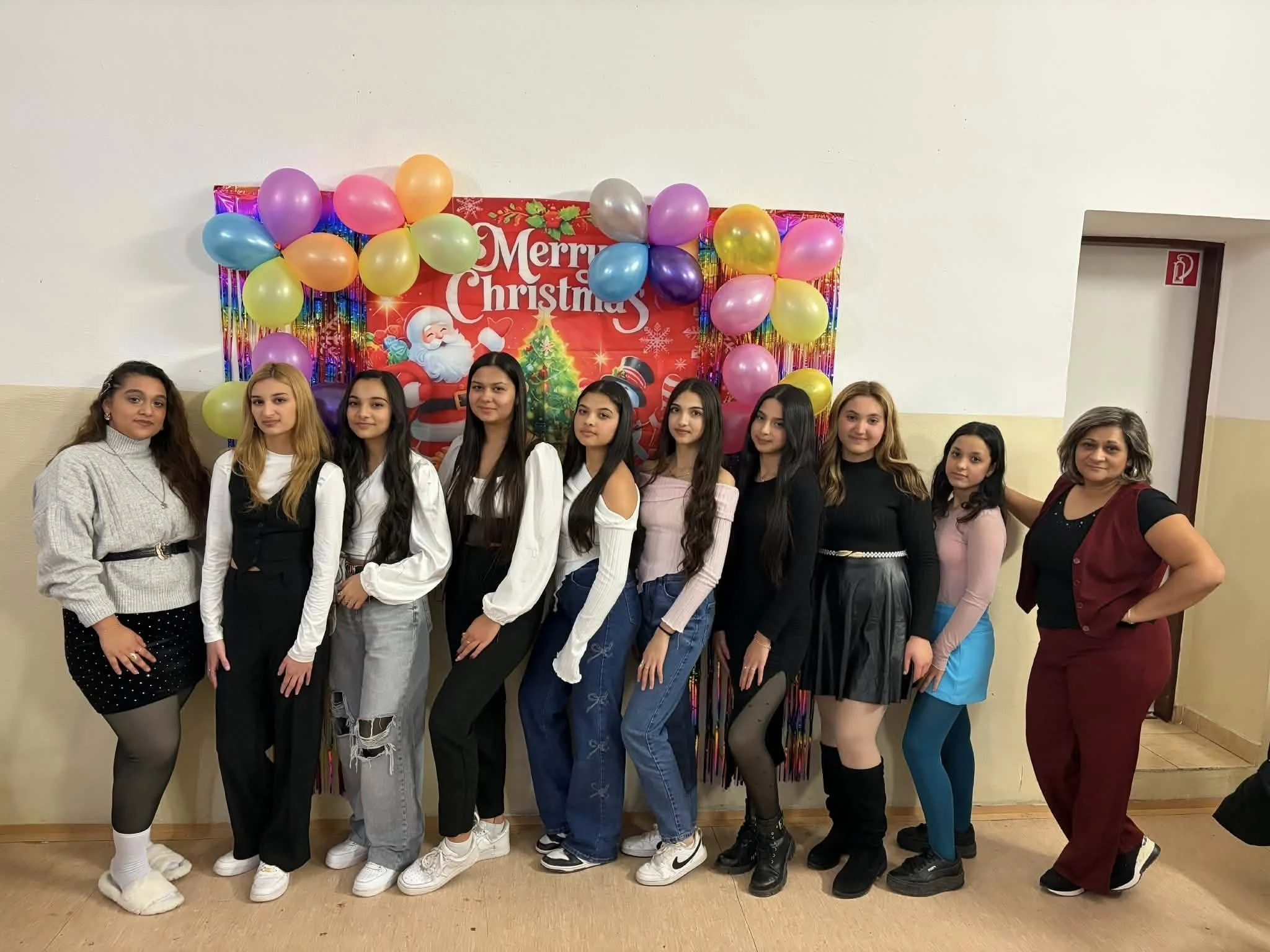 Group of young women and a woman teacher posing in front of a Christmas-themed backdrop with balloons and a "Merry Christmas" sign.