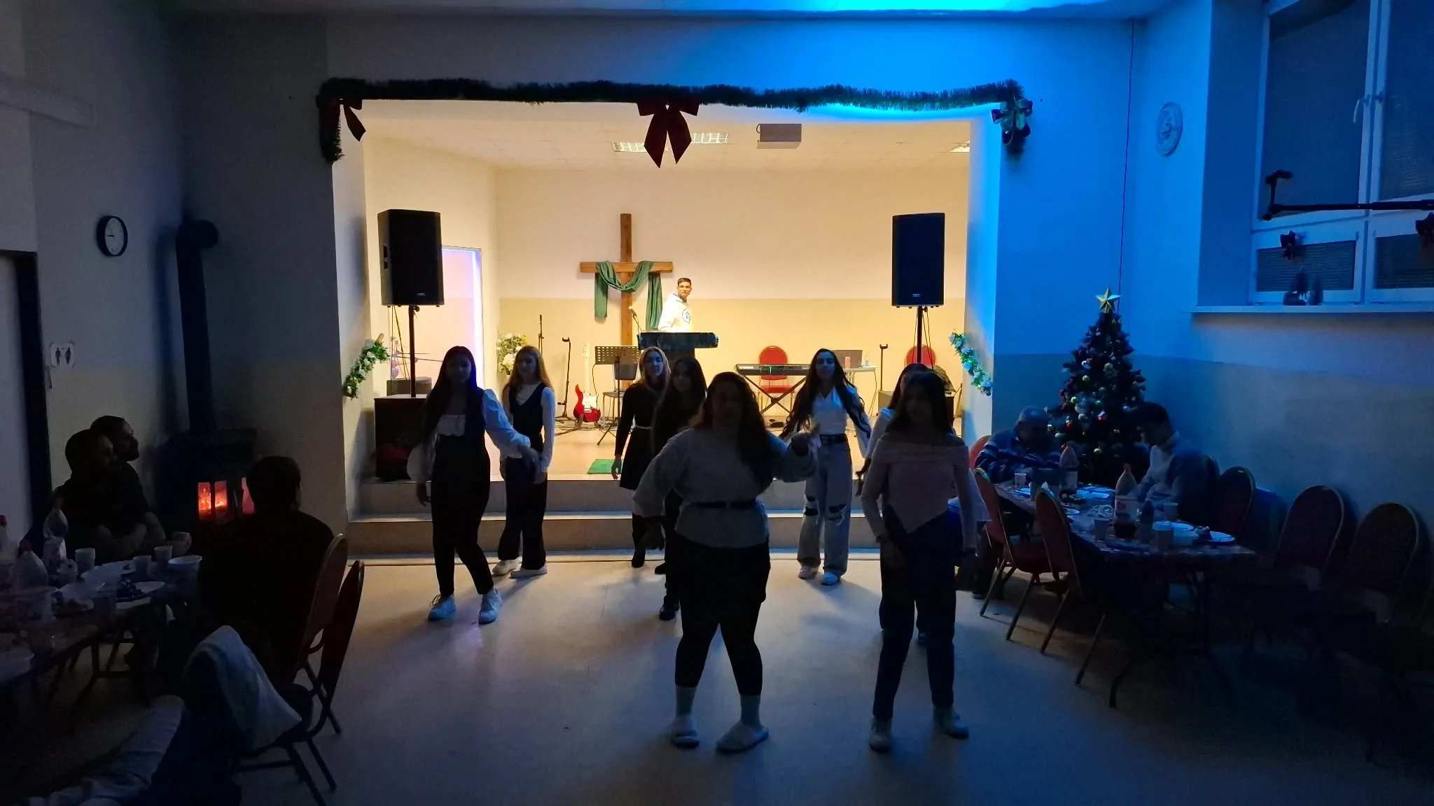 A group of people dancing in front of a decorated stage with Christmas ornaments and a Christmas tree on the side, in a room with tables and seated guests.