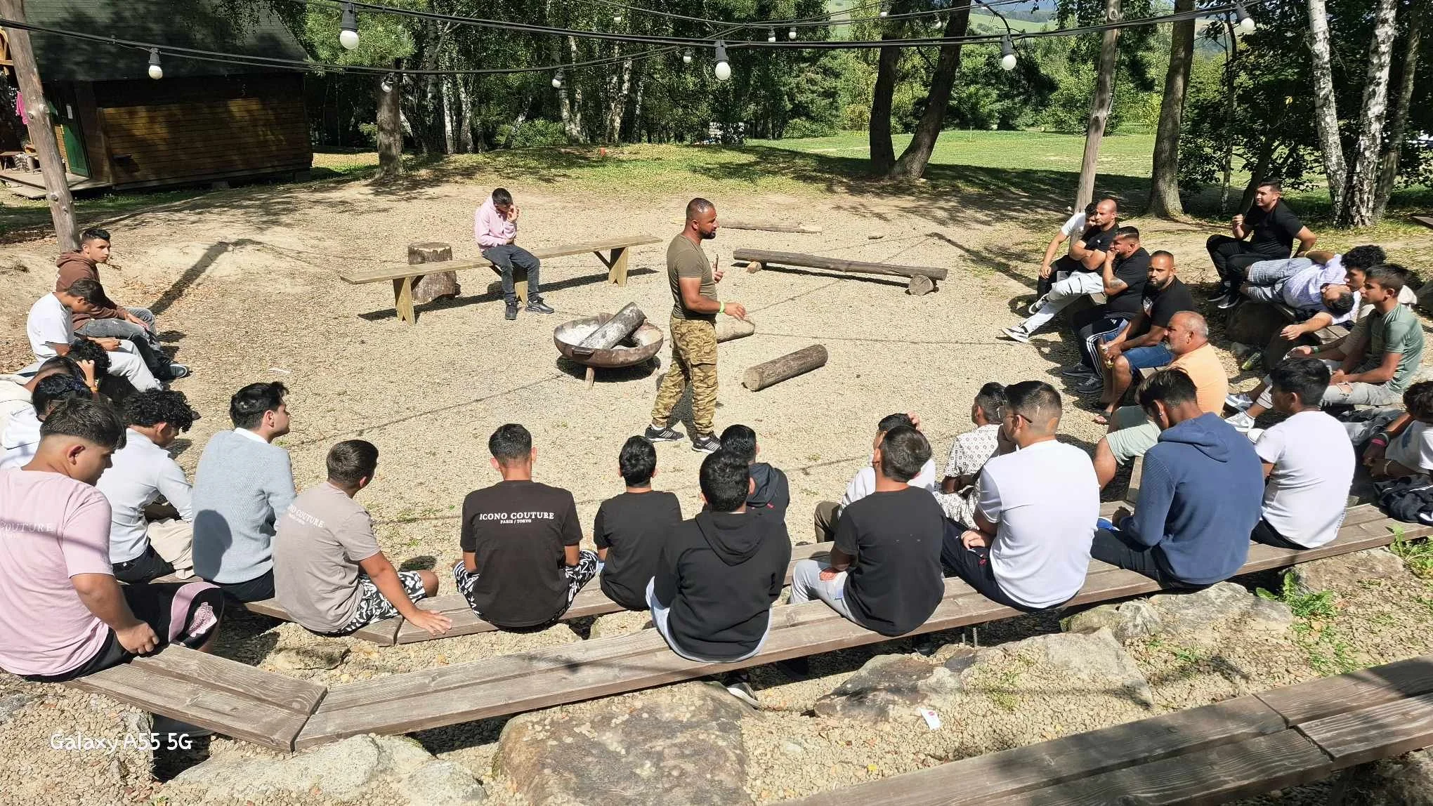 A group of young men sitting on wooden benches arranged in a semi-circle outdoors, listening to a man standing in the center who is giving a presentation or speech next to logs and a fire pit. There are trees and string lights overhead.
