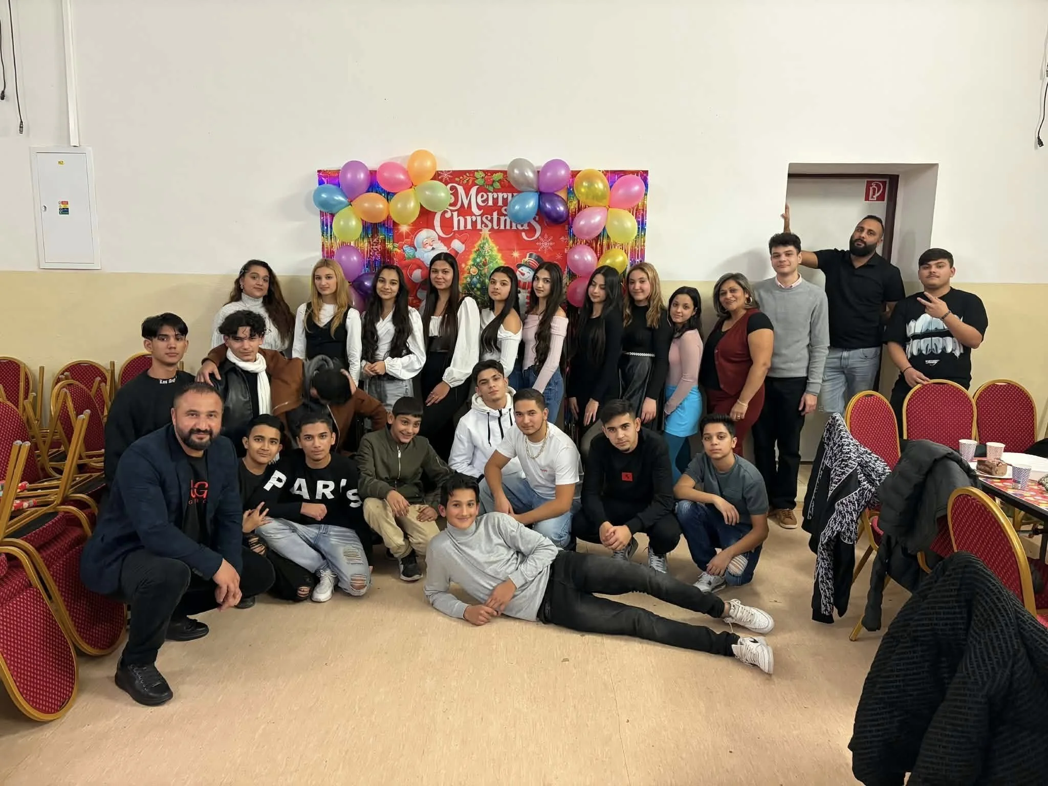 Group of young people and adults celebrating Christmas indoors with balloons and a decorated backdrop that says 'Merry Christmas'.