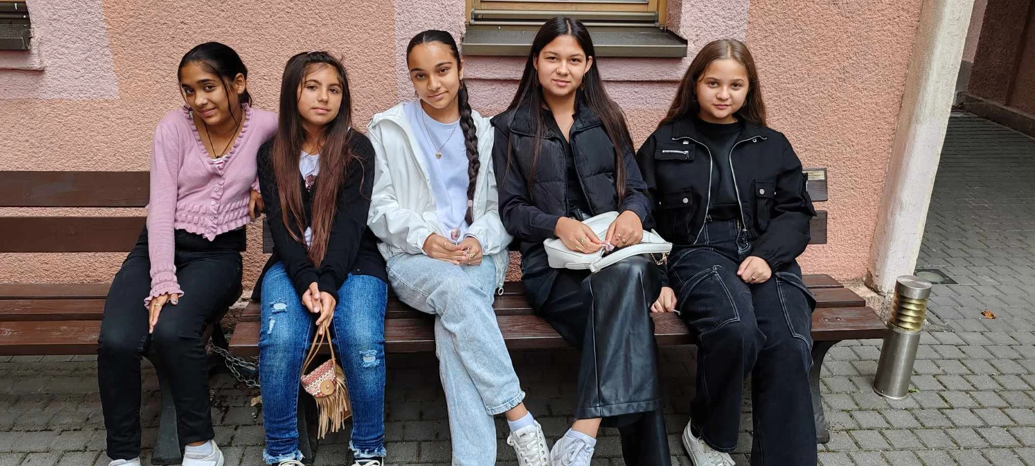 Five young girls sitting on a wooden bench outside against a pink textured wall. All are dressed casually and looking at the camera.