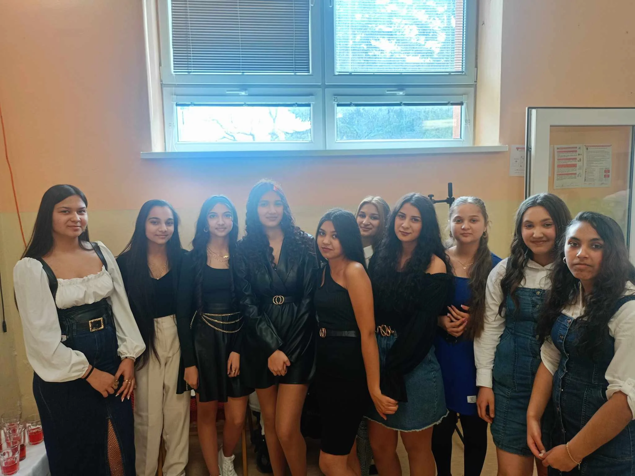 A group of eleven young women standing indoors in front of windows, dressed in various stylish outfits, smiling and posing for a photo.