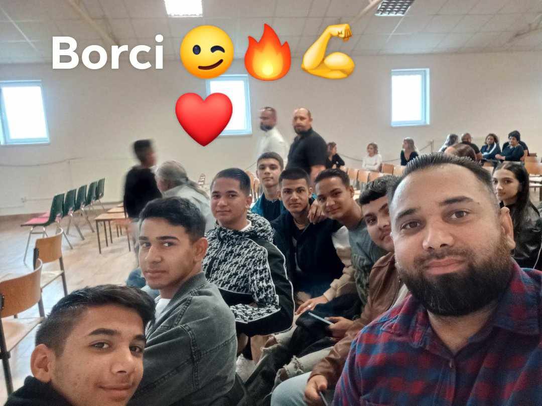 Group of young people and an adult man sitting in a classroom or auditorium, with some standing in the background, all facing the camera for a selfie.