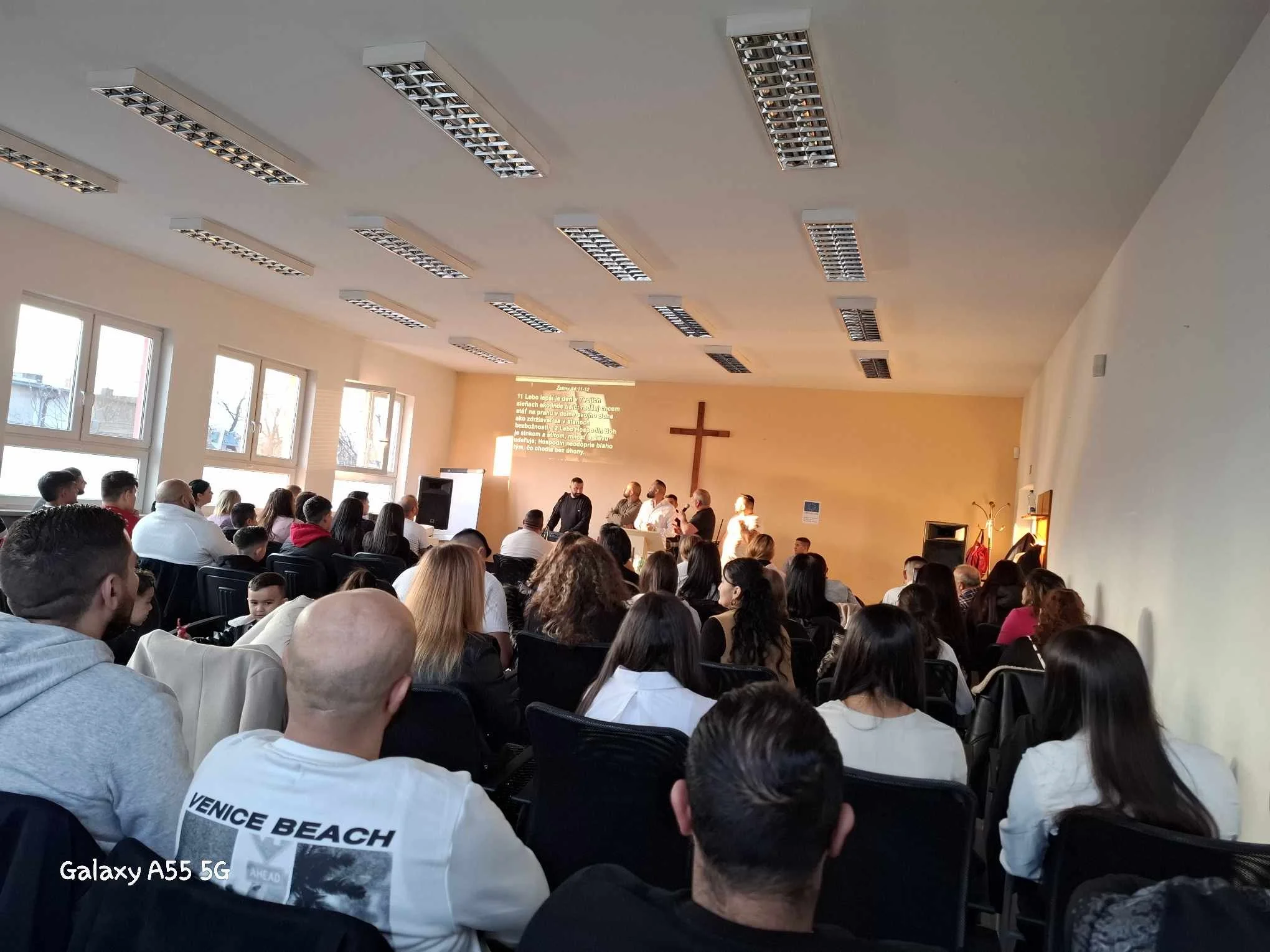 Inside a church or community hall with a congregation gathered, facing the front where a cross is mounted on the wall. Several people are singing or leading a song at the front, where a projection screen displays text. The room is well-lit with natur