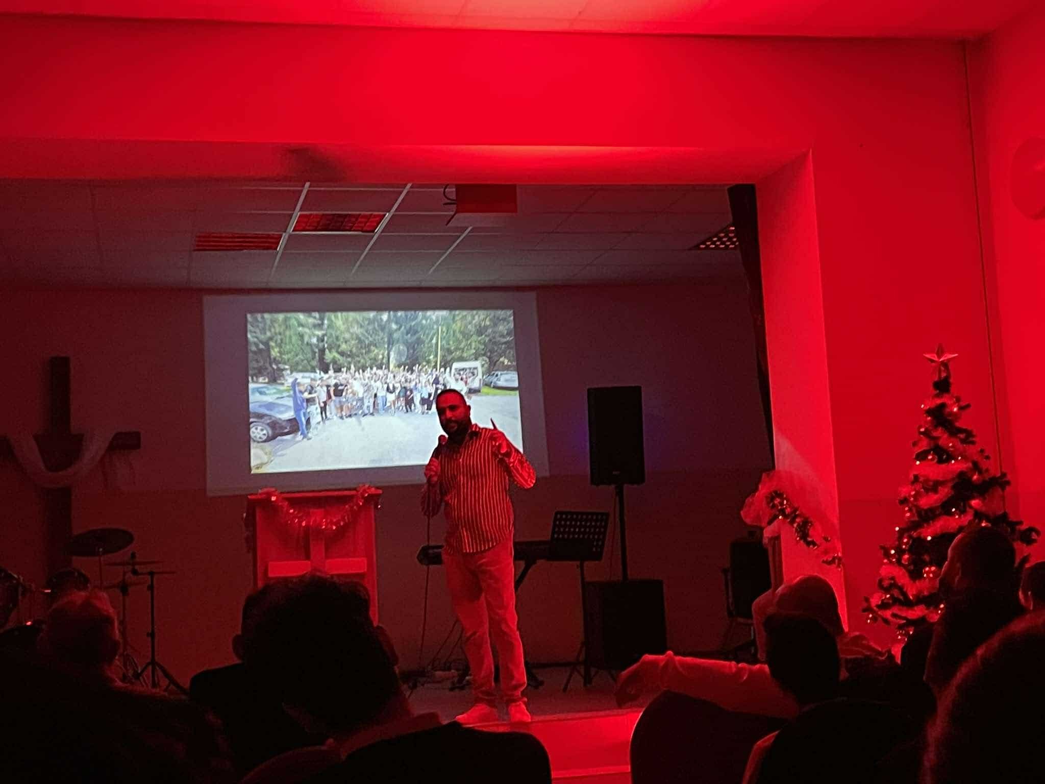 A man stands on stage in a dimly lit room with red lighting, holding a microphone and smiling. Behind him, a large screen displays a photo of a crowd gathered outdoors. The stage is decorated with a Christmas tree and festive garland.
