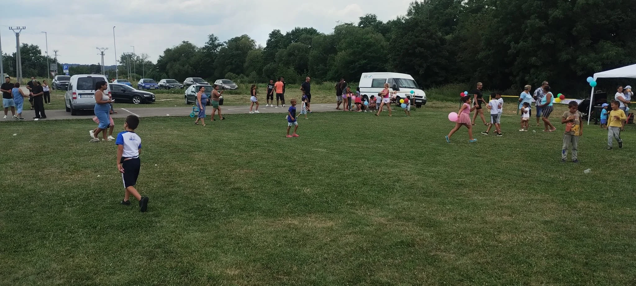 Children playing on a grassy field at a community gathering, with some holding balloons, and adults standing or walking nearby, a row of parked cars and trees in the background, and a white tent with colorful balloons on the right.
