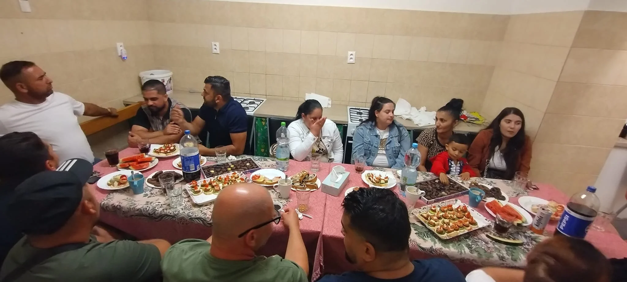 A group of people gathered around a table filled with food and desserts at a celebration or party in an indoor setting.