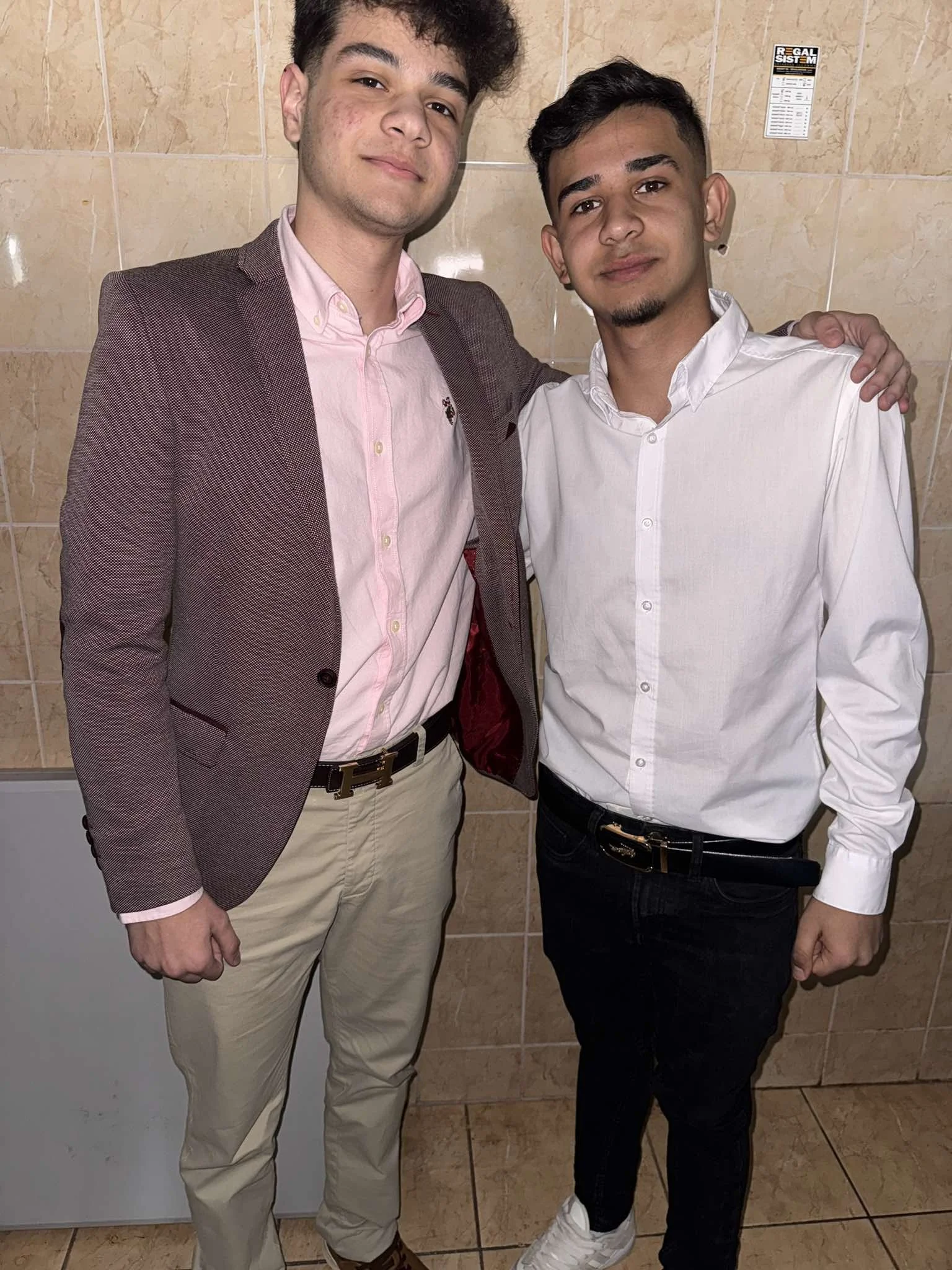 Two young men dressed in formal attire standing close together, one with a light pink shirt and blazer, the other with a white shirt, in front of a tiled wall.