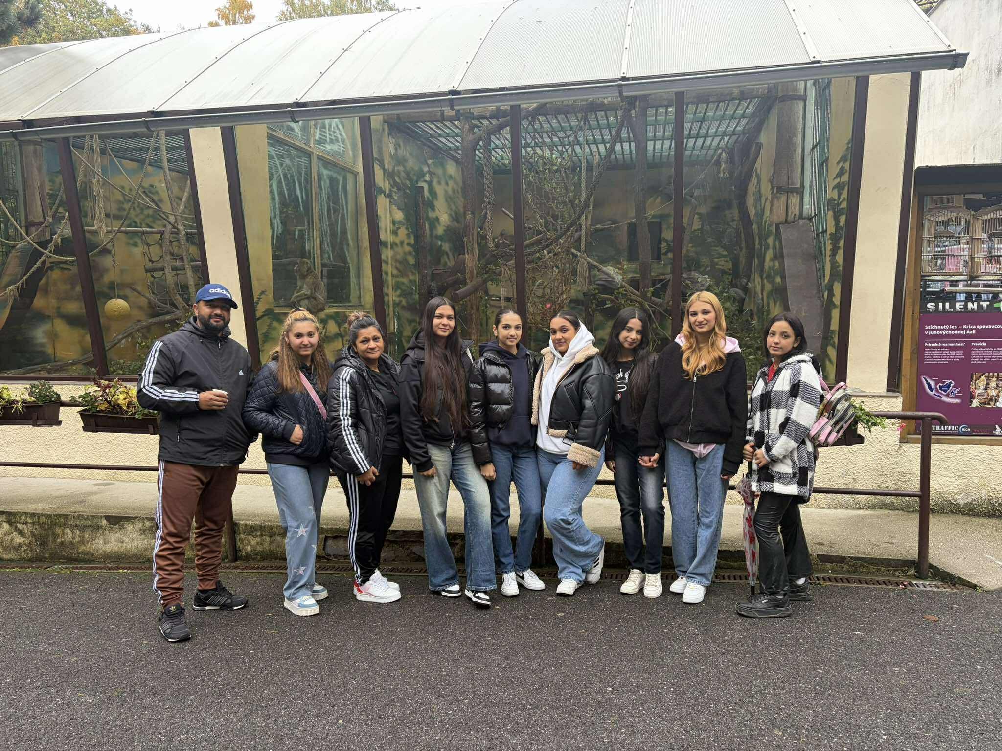 Group of nine people standing in front of glass enclosure with trees and animals inside at a zoo or wildlife park.