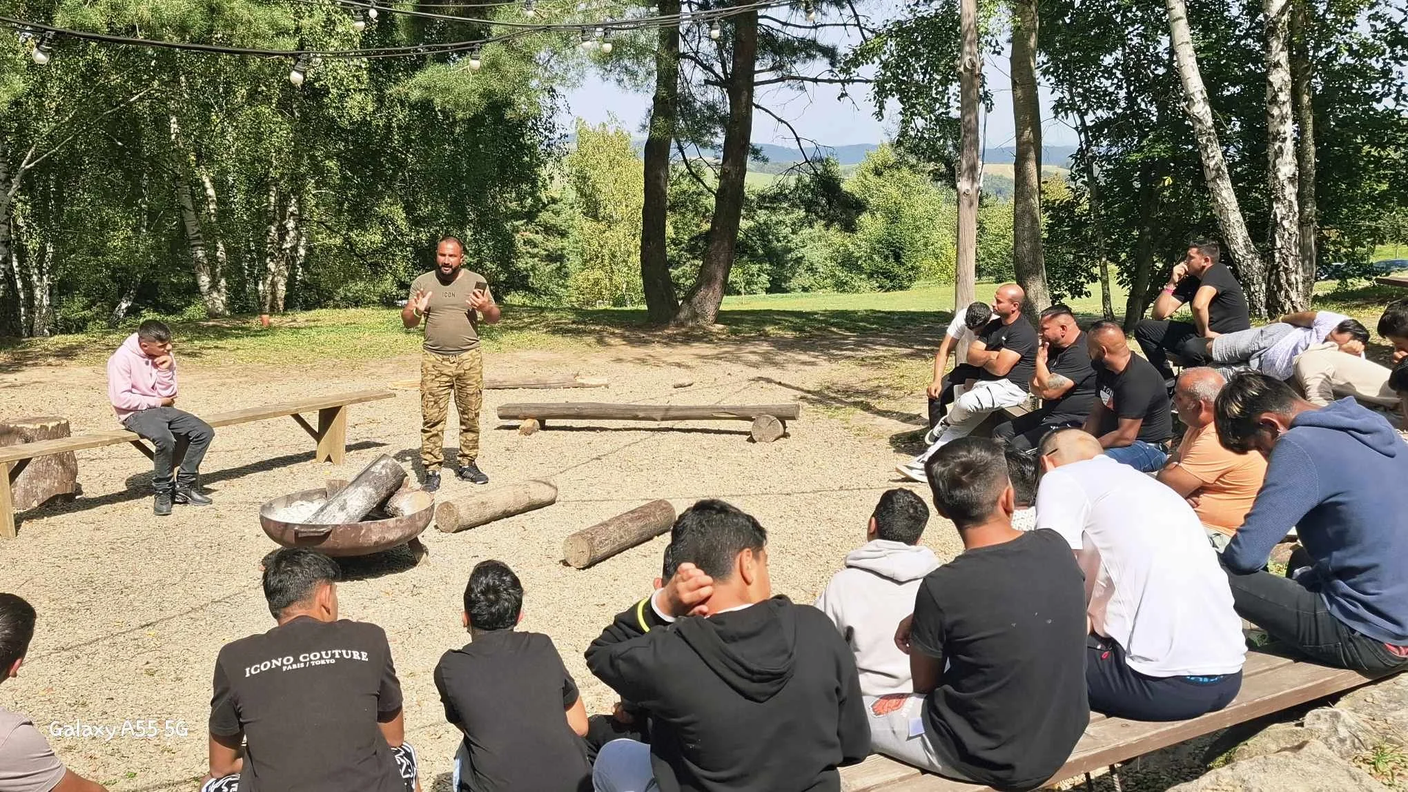 A group of people sitting outdoors on a wooden bench and logs, listening to a man speaking at an outdoor event surrounded by trees.