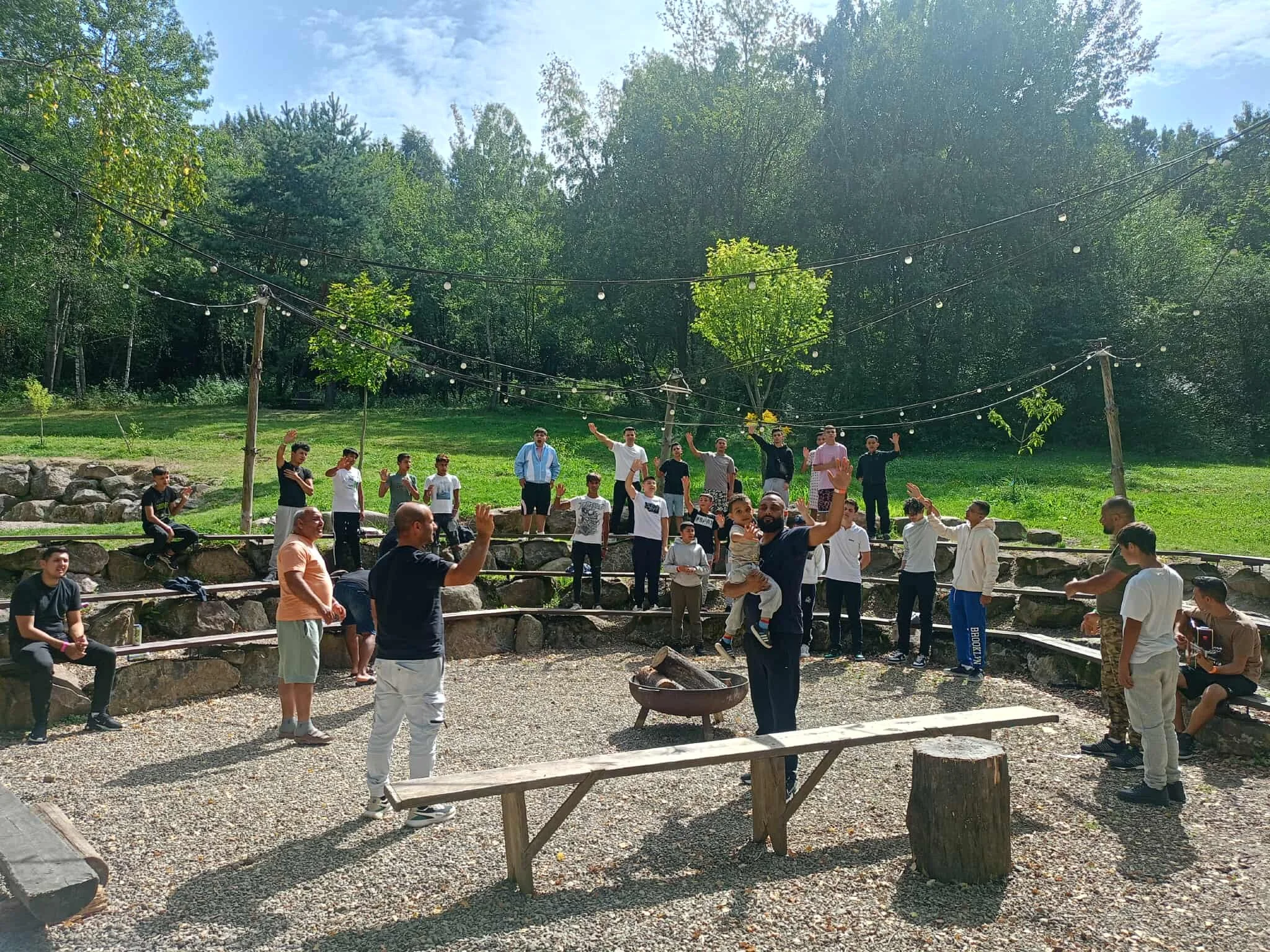 A group of children and adults gathered outdoors in a circle, some standing on tiered rocks, under string lights, with trees and a grassy hill in the background, during daytime.